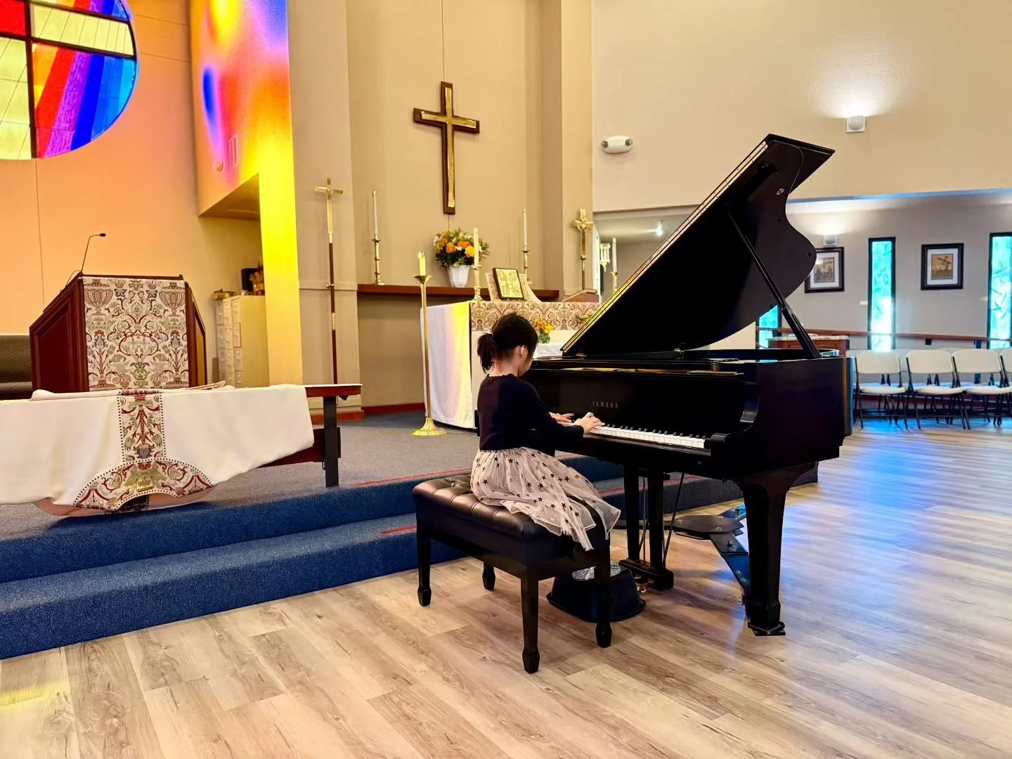 A young girl in a black top and a beige skirt with star patterns playing a black Yamaha grand piano in a church, with religious symbols and stained glass windows in the background.