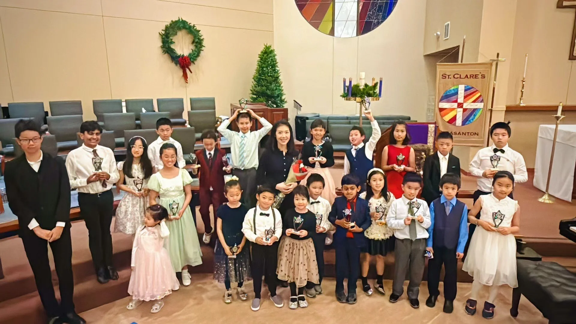 Group of children and adults dressed in formal attire, holding trophies, in a church or hall decorated with a Christmas wreath and a small Christmas tree.