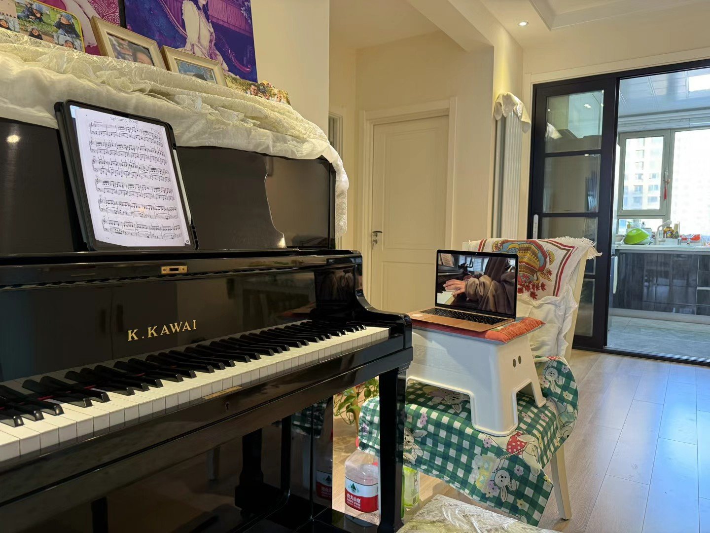 A black K. Kawai grand piano with sheet music on its music stand in a well-lit room. There is a laptop on a white chair covered with a colorful cloth, showing a person playing piano. In the background, a balcony with sliding glass doors reveals an outdoor area. The room has wooden flooring and decorated walls with pictures and art.