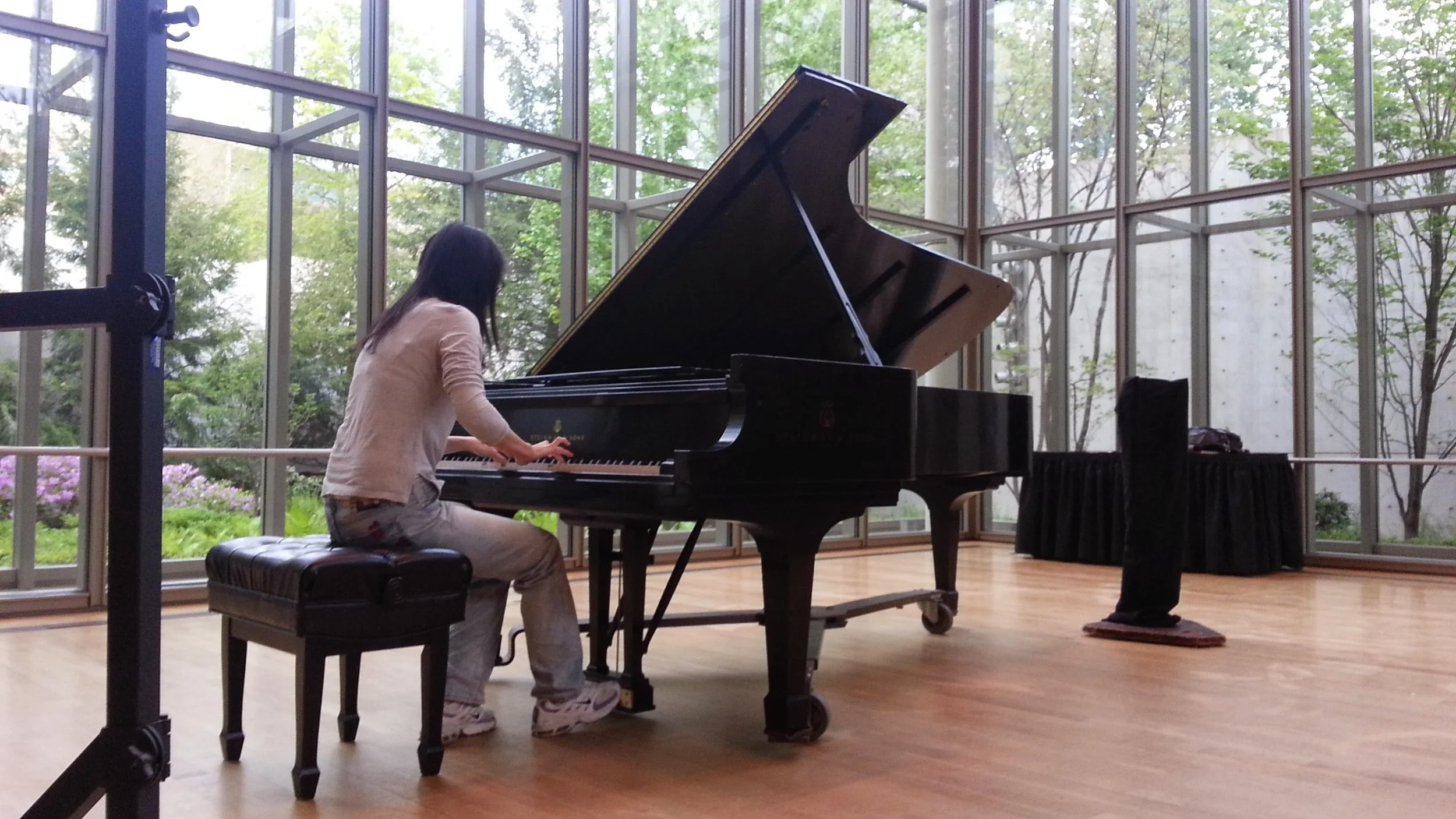 A person sitting on a bench playing a grand piano in a bright, spacious room with large glass windows and a view of trees outside.