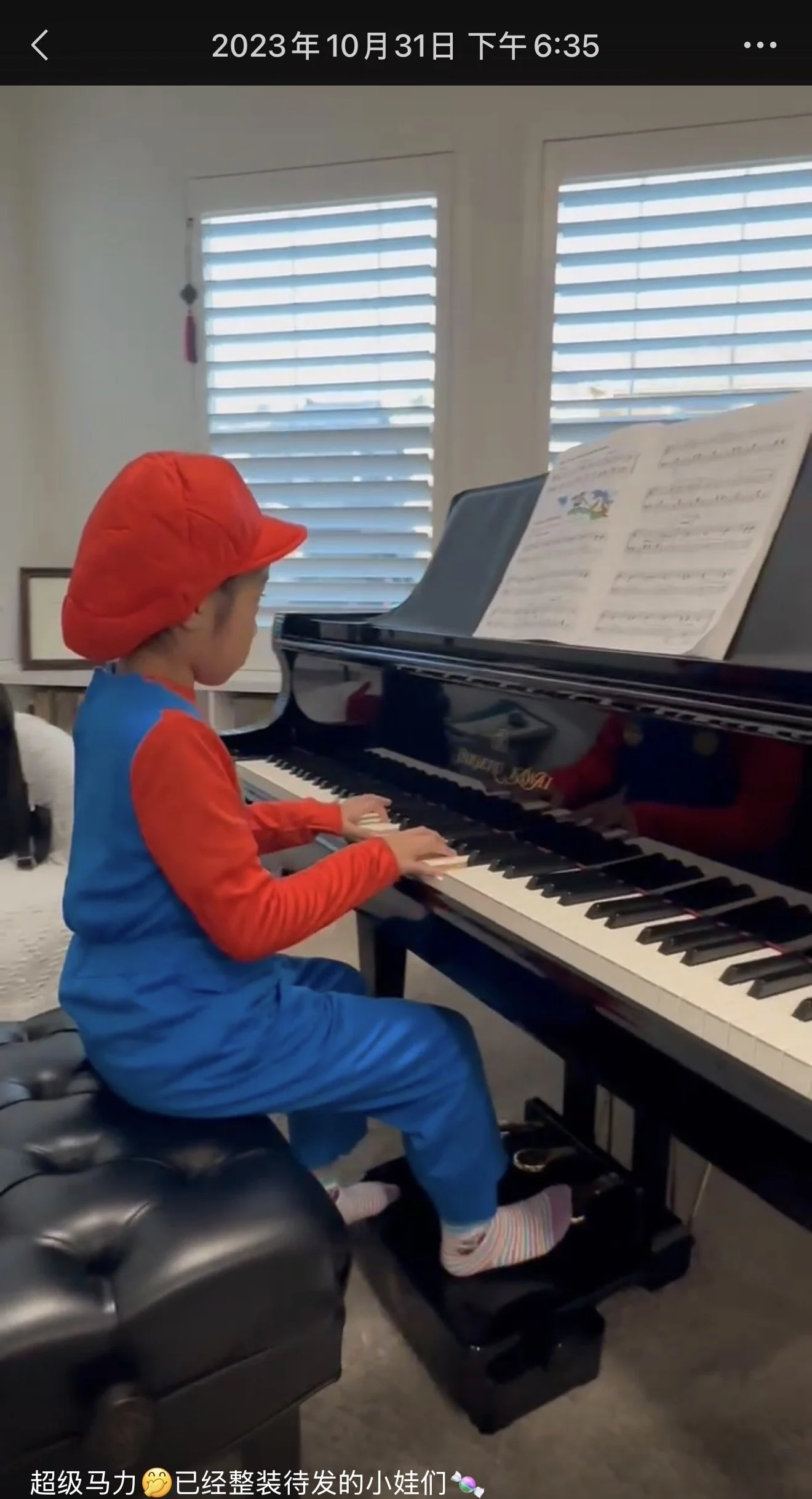 A young child dressed in a colorful costume, playing a grand piano in a bright room with windows and blinds.