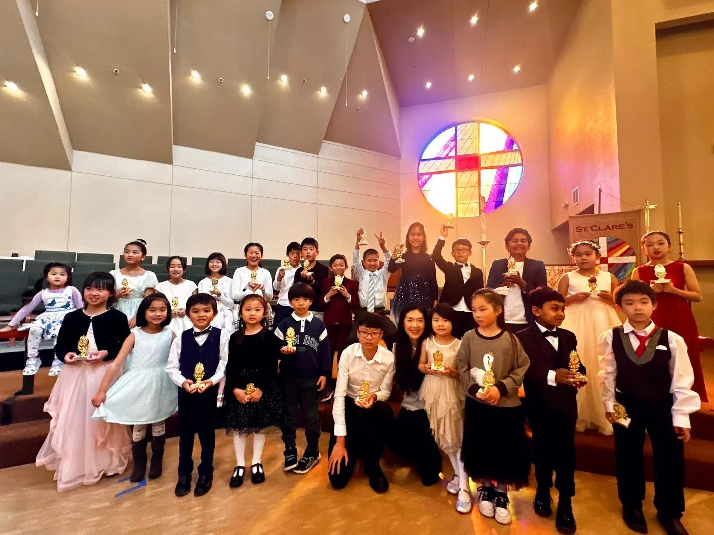 Group of children and adults in formal attire holding trophies inside a church with stained glass window and cross