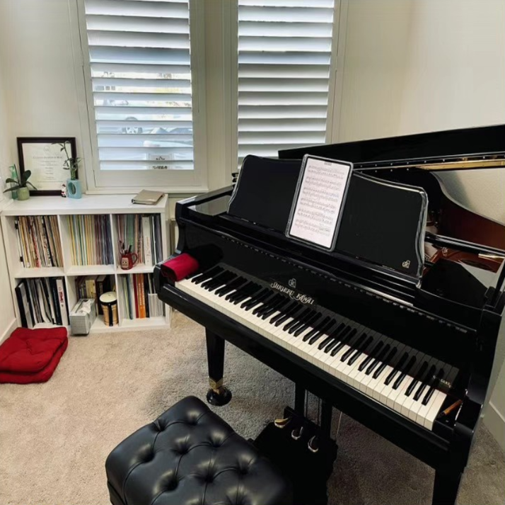 A black grand piano with a sheet music tablet on its music stand in a room with beige carpeting and a white bookshelf filled with books and vinyl records. There is a window with white shutters behind the piano, and a red cushion is on the floor next to the bookshelf.
