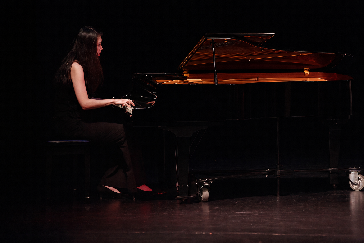 A woman in black attire playing a grand piano on a dark stage.