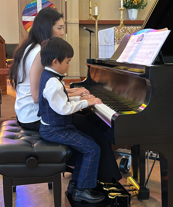 A young boy and woman playing piano together in a church or music room, with sheet music on the piano, a colorful stained glass window in the background, and religious candles and decor.