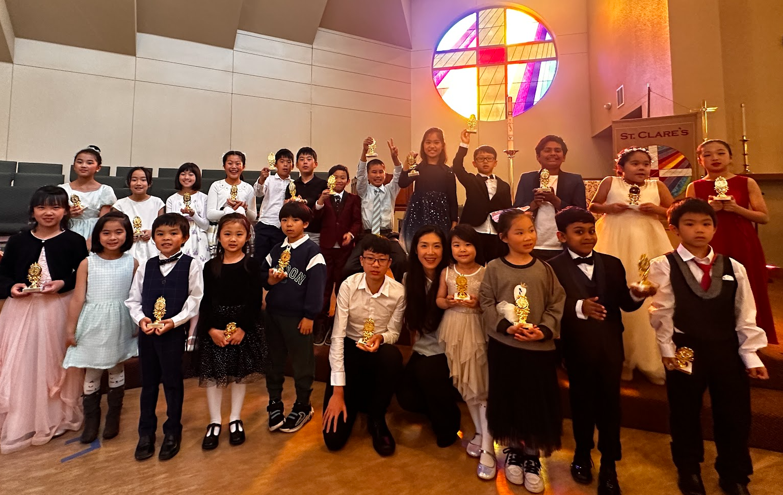 Group of children and a woman in a church, holding awards, with a stained glass window in the background, celebrating a special event or ceremony.
