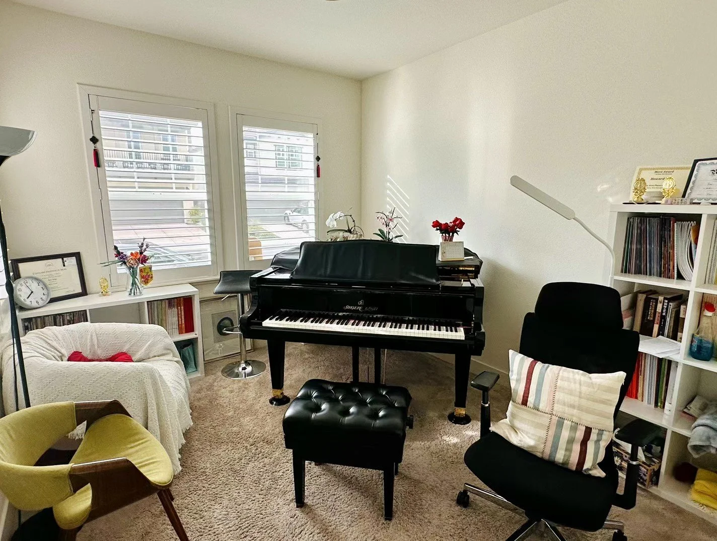 A music room with a black grand piano, an ergonomic black office chair with a striped pillow, a white bookshelf with books and awards, a beige armchair with red pillows, a yellow armchair, and a white console table with decorative items. Two windows with blinds are behind the piano.