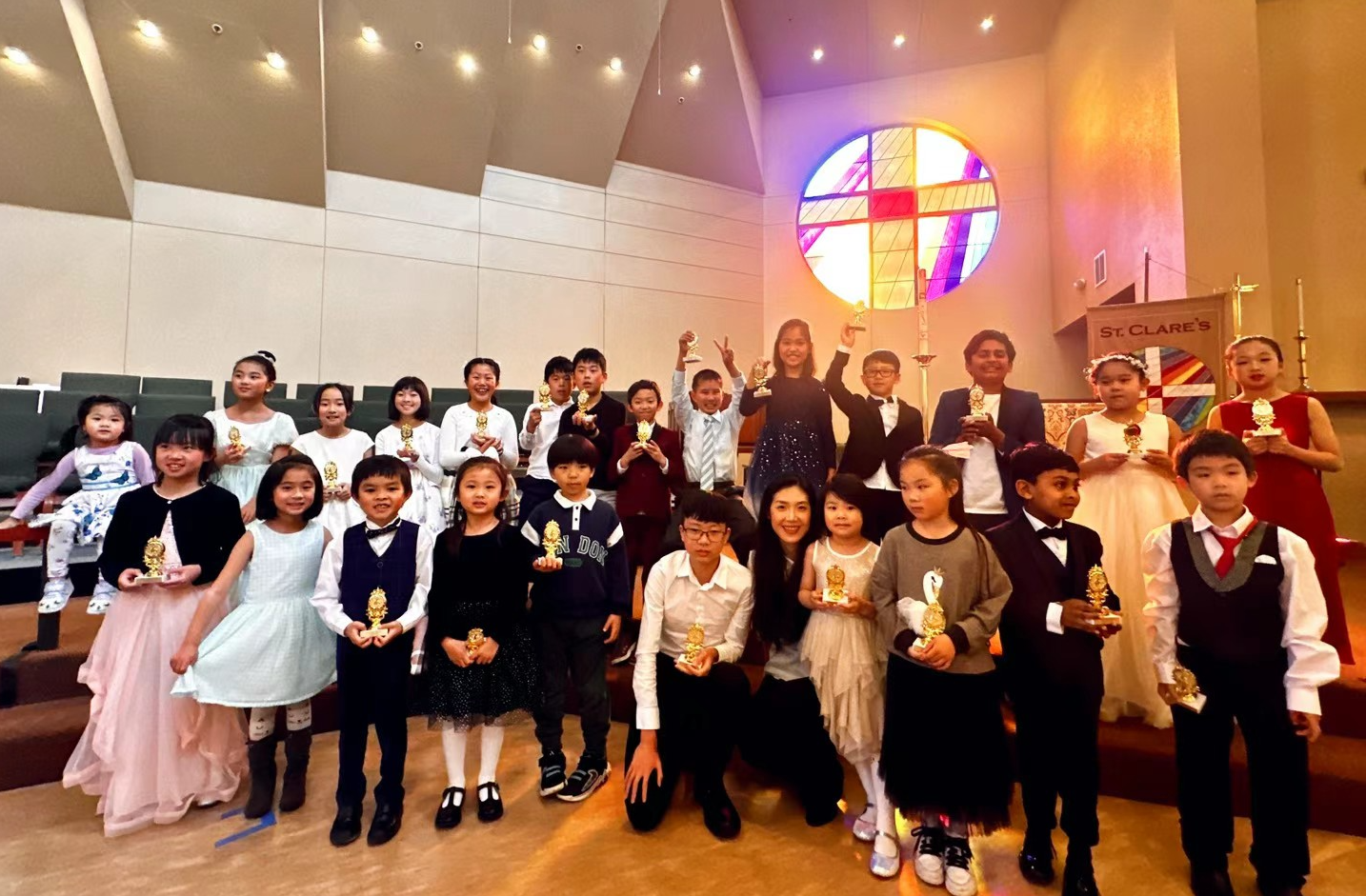 Children and adults dressed in formal attire standing on church steps, holding trophies after a ceremony, with a stained glass cross window in the background.