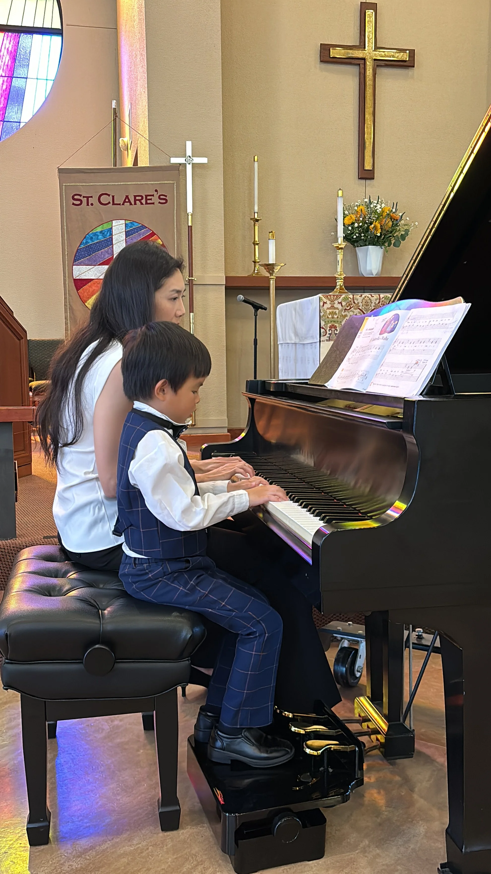 A woman and a young boy playing the piano together inside a church, with religious symbols and flowers in the background.
