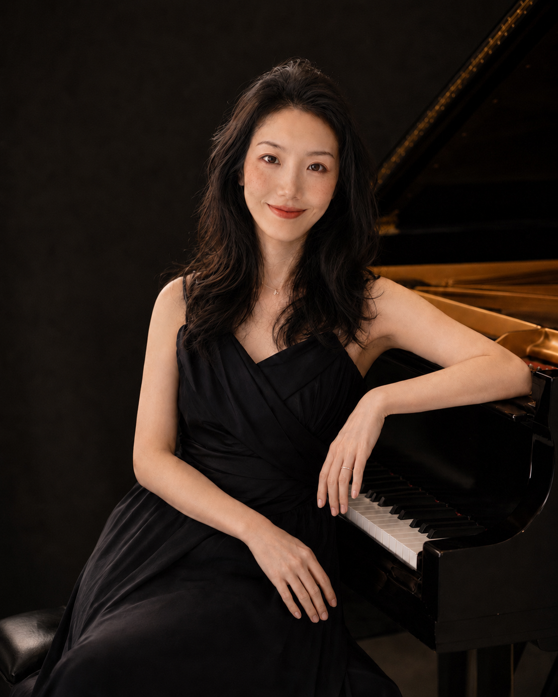 A young woman in a black dress sitting at a grand piano, smiling gently at the camera.