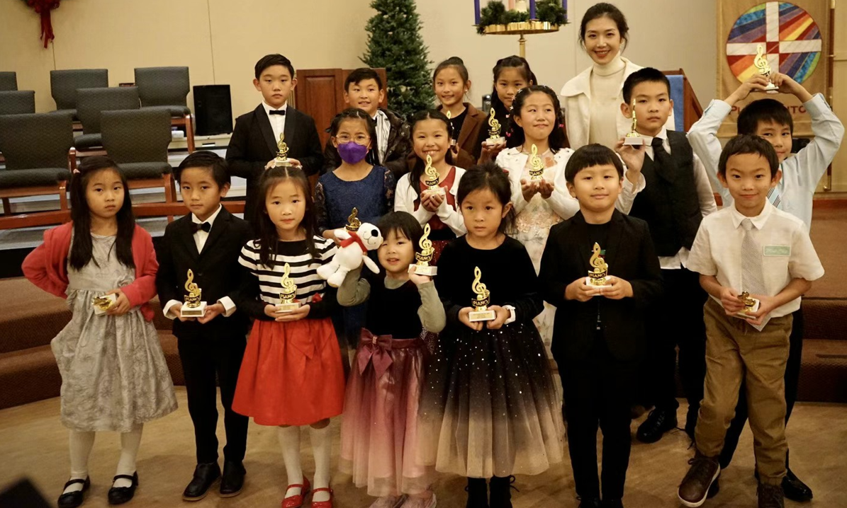 Children in formal attire holding awards and trophies, posing with a woman on stage in a decorated room.
