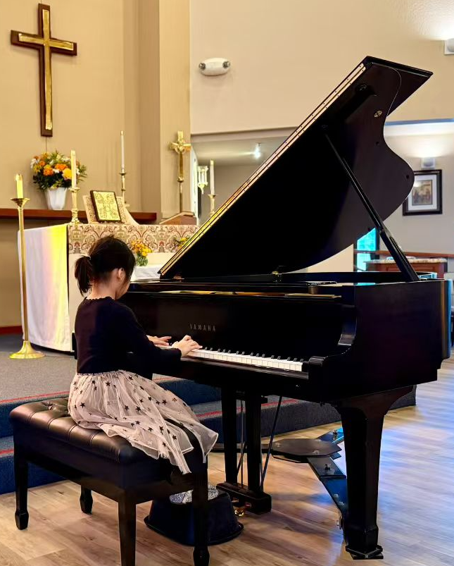 A young girl sitting on a piano bench playing a black Yamaha grand piano in a church or chapel, with religious symbols and flowers in the background.