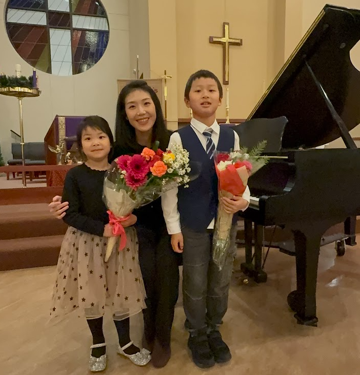 A woman and two children standing inside a church, holding flowers, with a grand piano and a cross in the background.