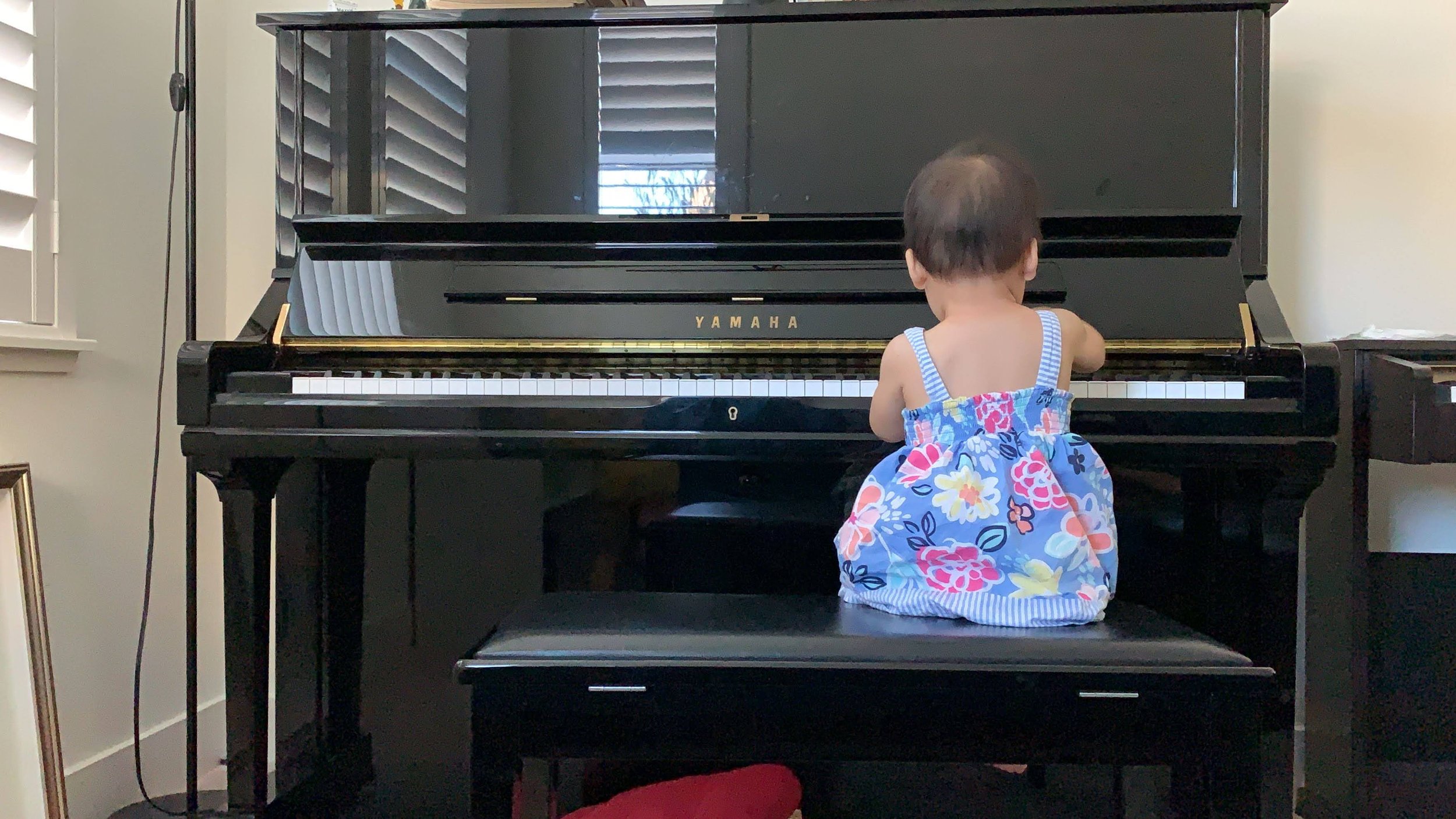 A young child sitting on a black piano bench, facing a black Yamaha upright piano, wearing a colorful floral dress.
