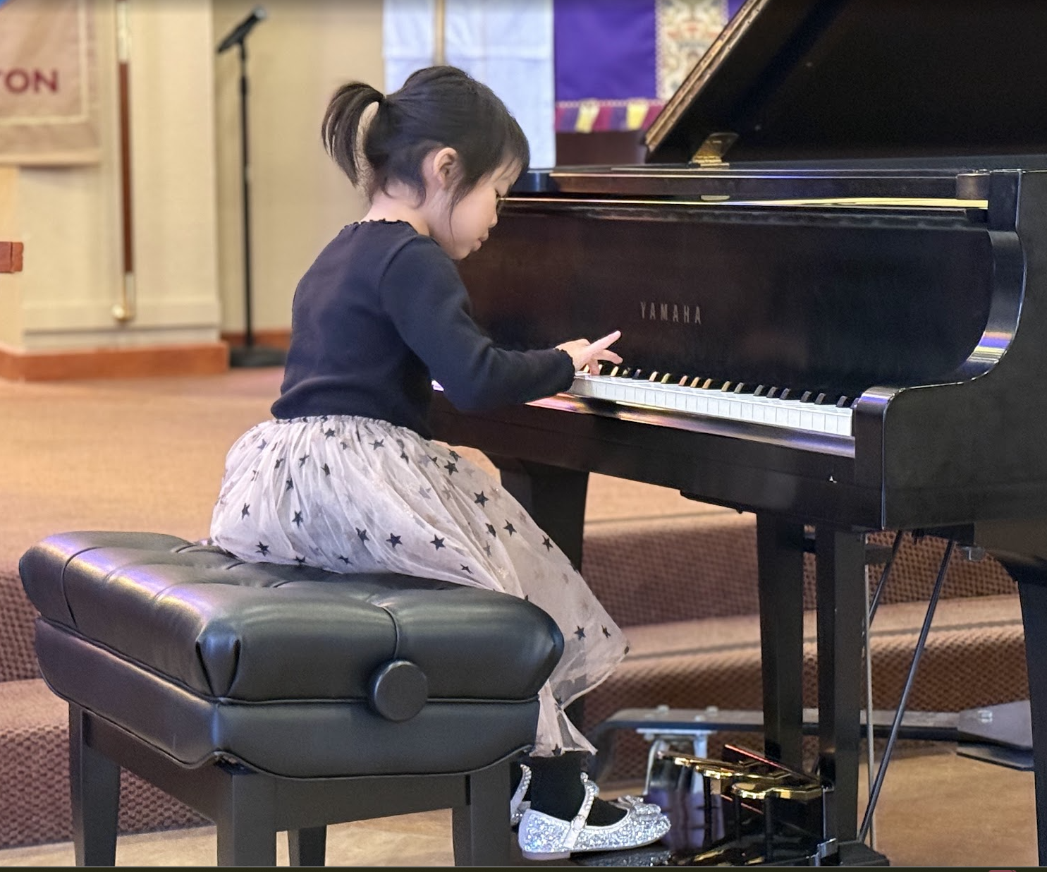 A young girl playing a black Yamaha grand piano, sitting on a black padded bench, wearing a black top, a beige skirt with black star patterns, and glittery silver shoes.