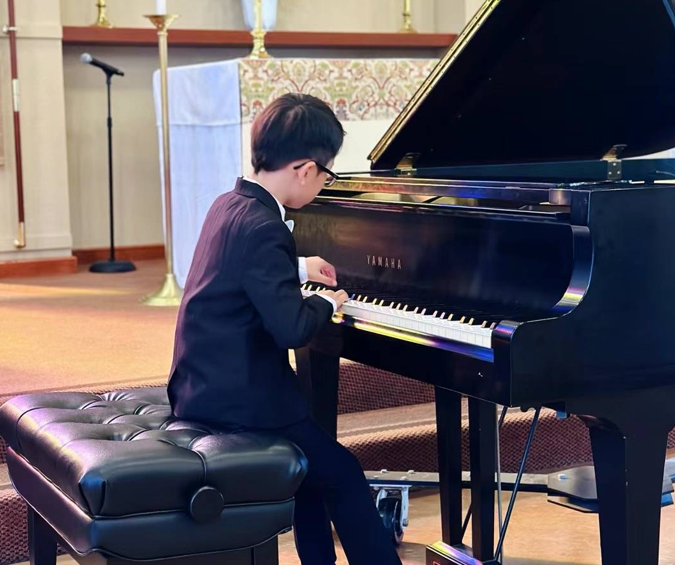 Young boy in a tuxedo playing a black Yamaha grand piano in a church or music hall setting.