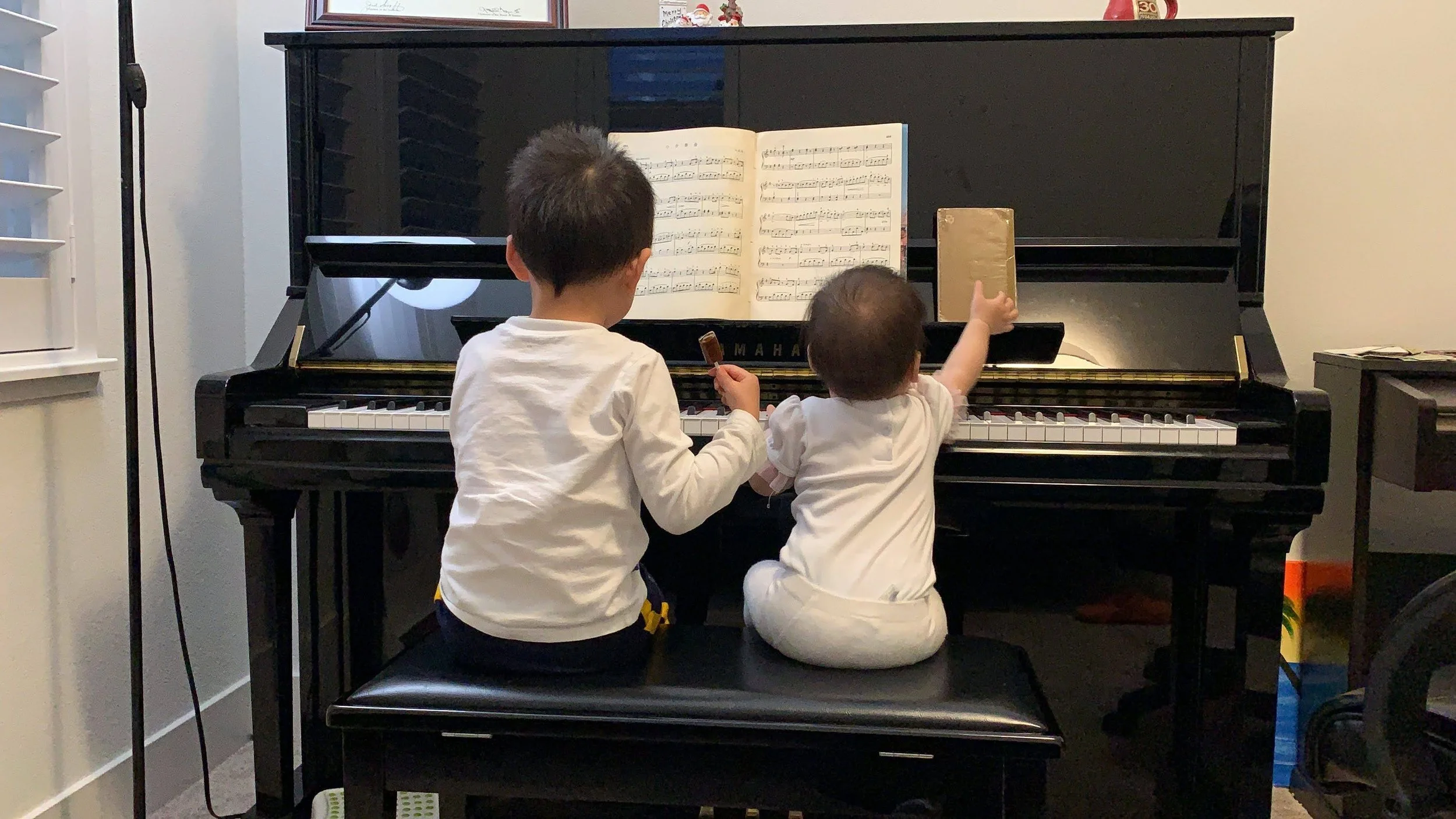 Two young children, one boy and one girl, sitting on a bench and playing a black Yamaha piano together in a room, with music sheet on the piano's music stand.
