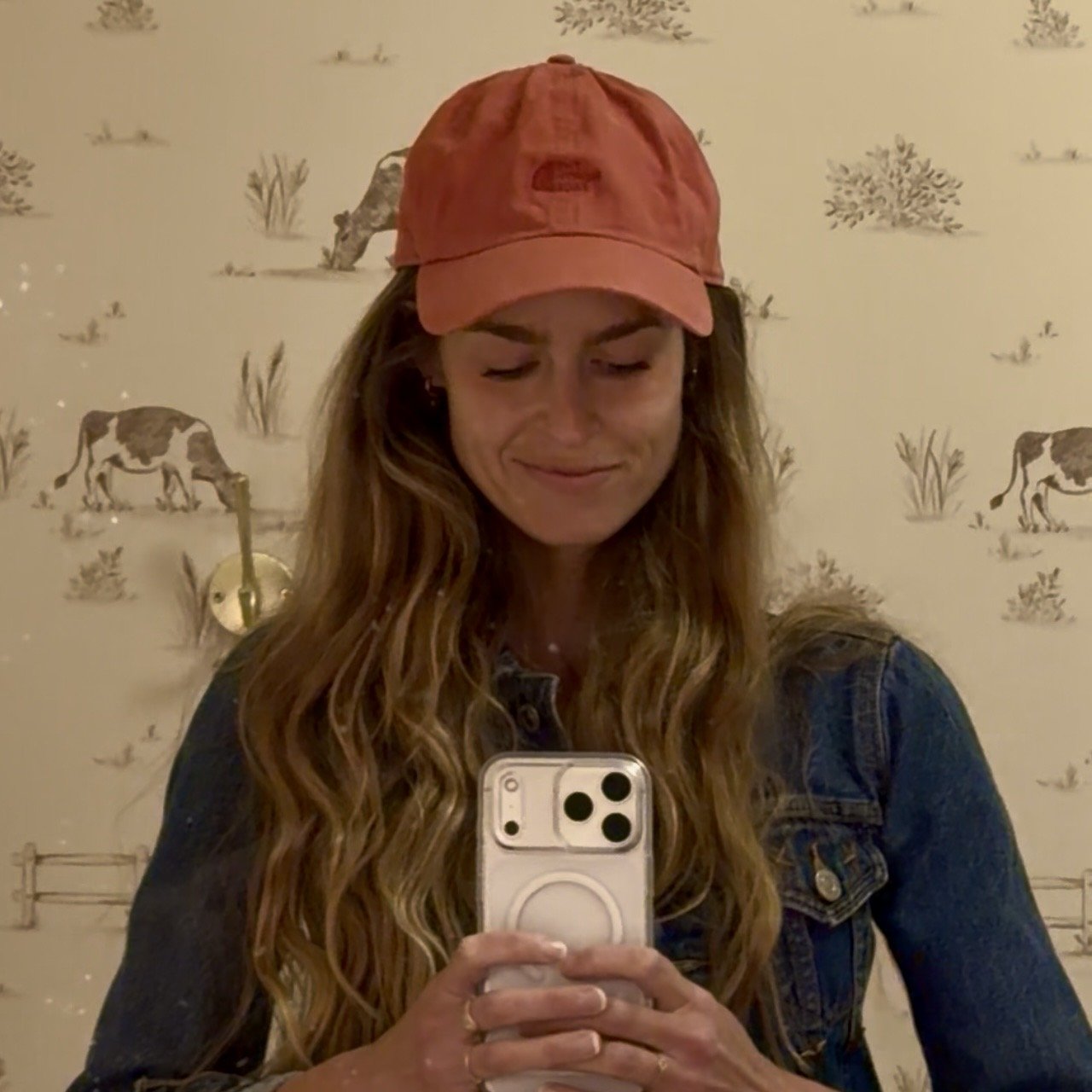 A woman with long, wavy hair wearing a pink baseball cap and a denim jacket, taking a selfie in front of a wallpaper with wildlife illustrations.