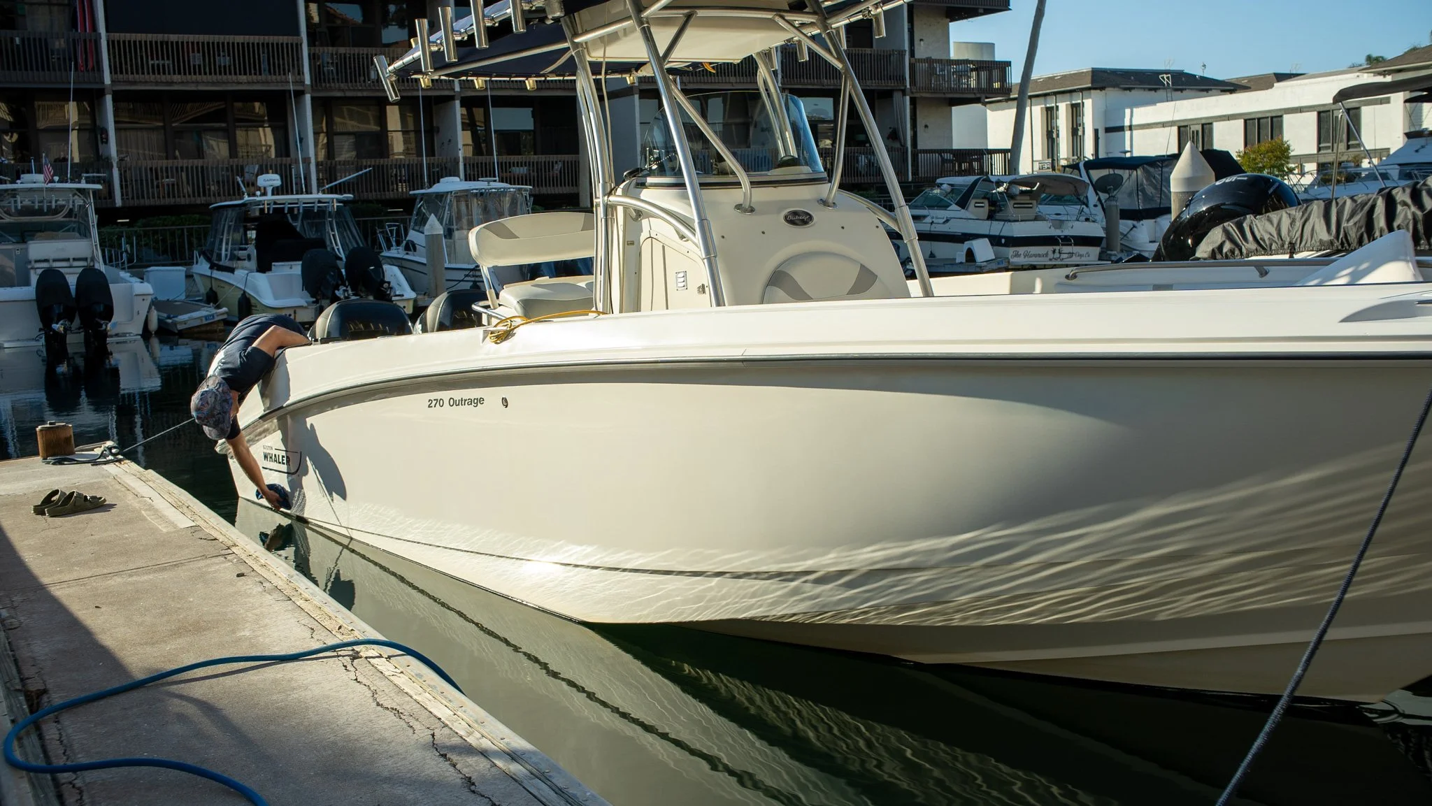 A person cleaning the side of a white boat named '270 Outrage' docked at a marina, with other boats and a building in the background.