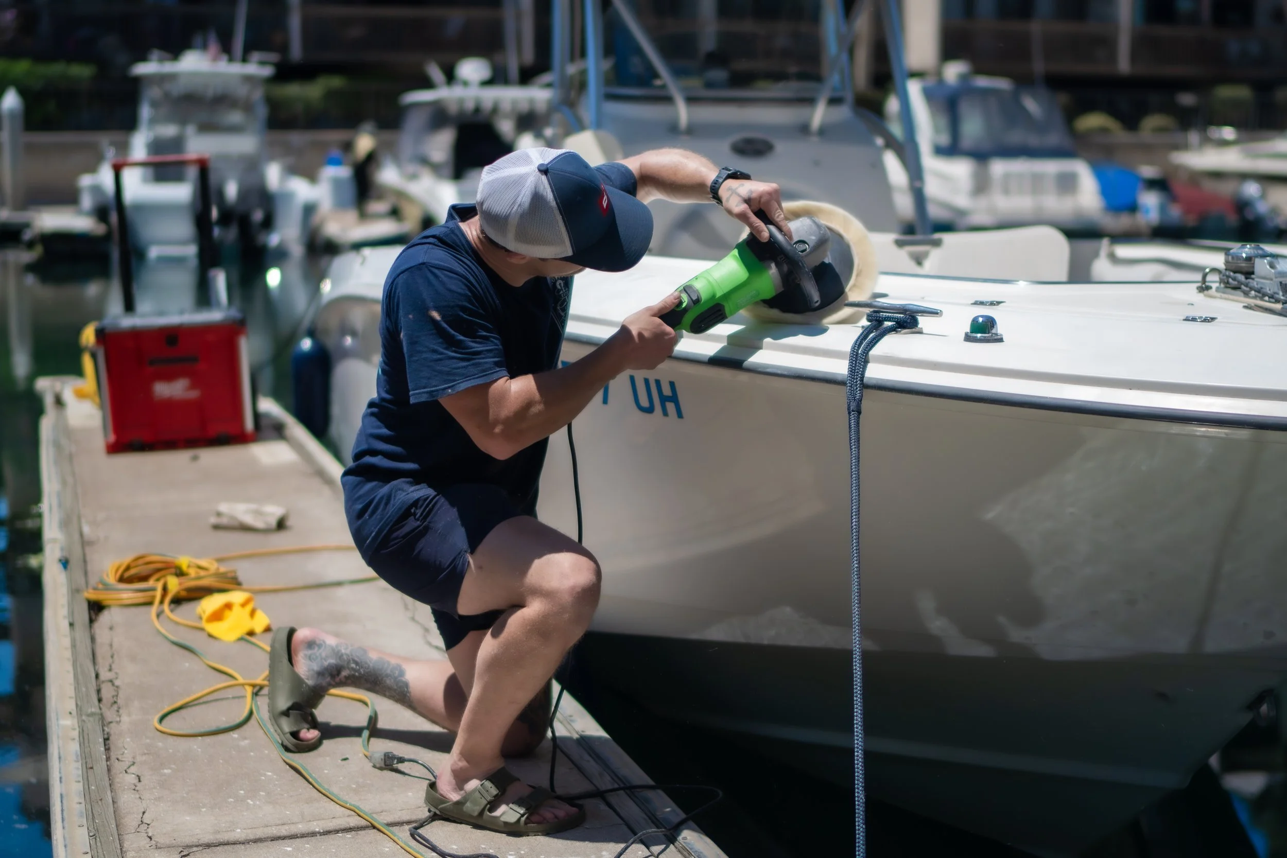A man kneeling on a dock, sanding the hull of a boat with a power sander. There are multiple boats docked in the background and various tools and cords on the dock.