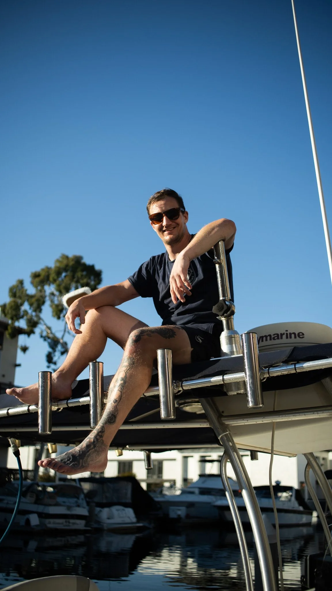 A young man with sunglasses sitting on the edge of a boat at a marina, smiling at the camera with a clear blue sky in the background.