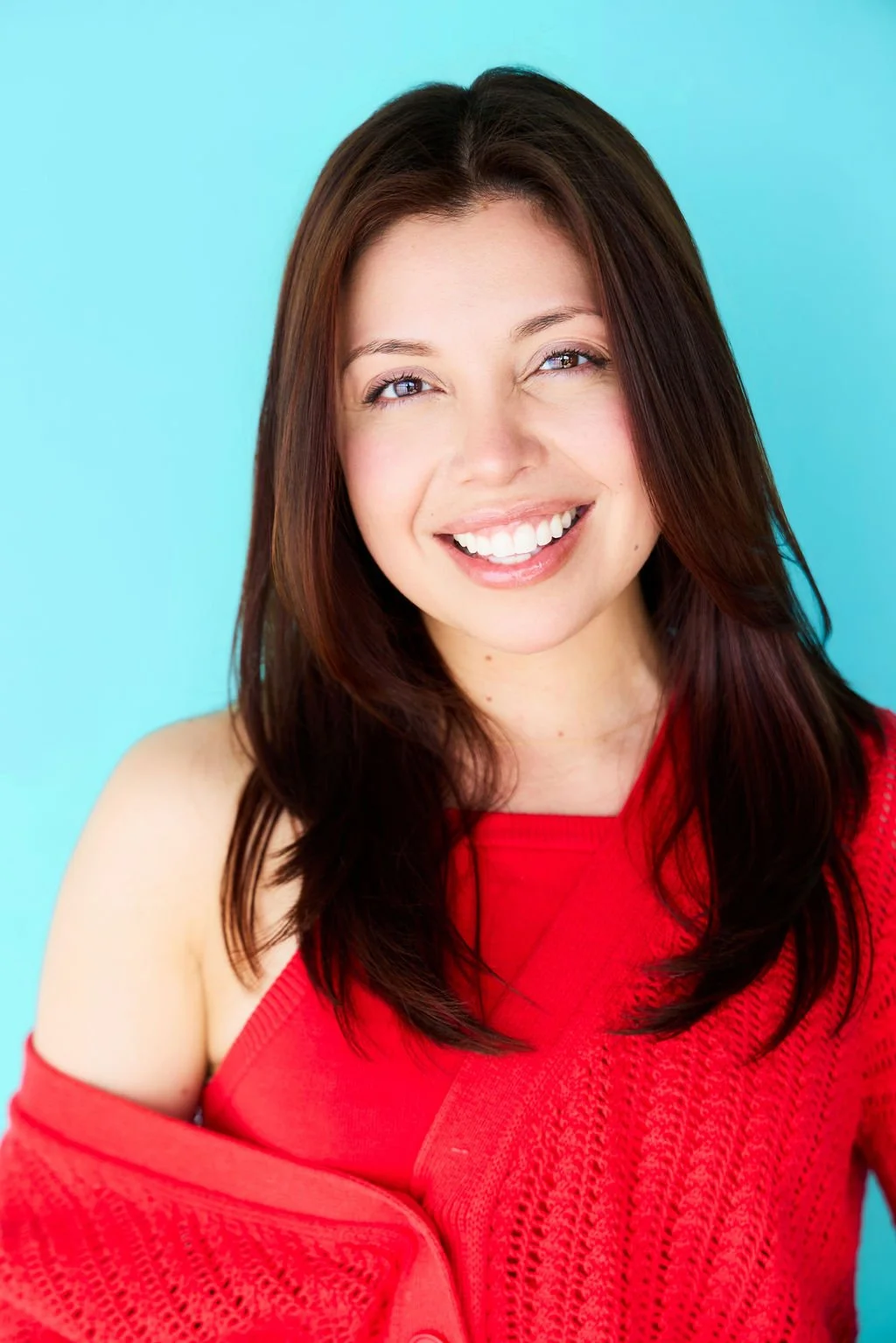 A woman with long brown hair smiling, wearing a red top and a matching red cardigan, against a bright blue background.