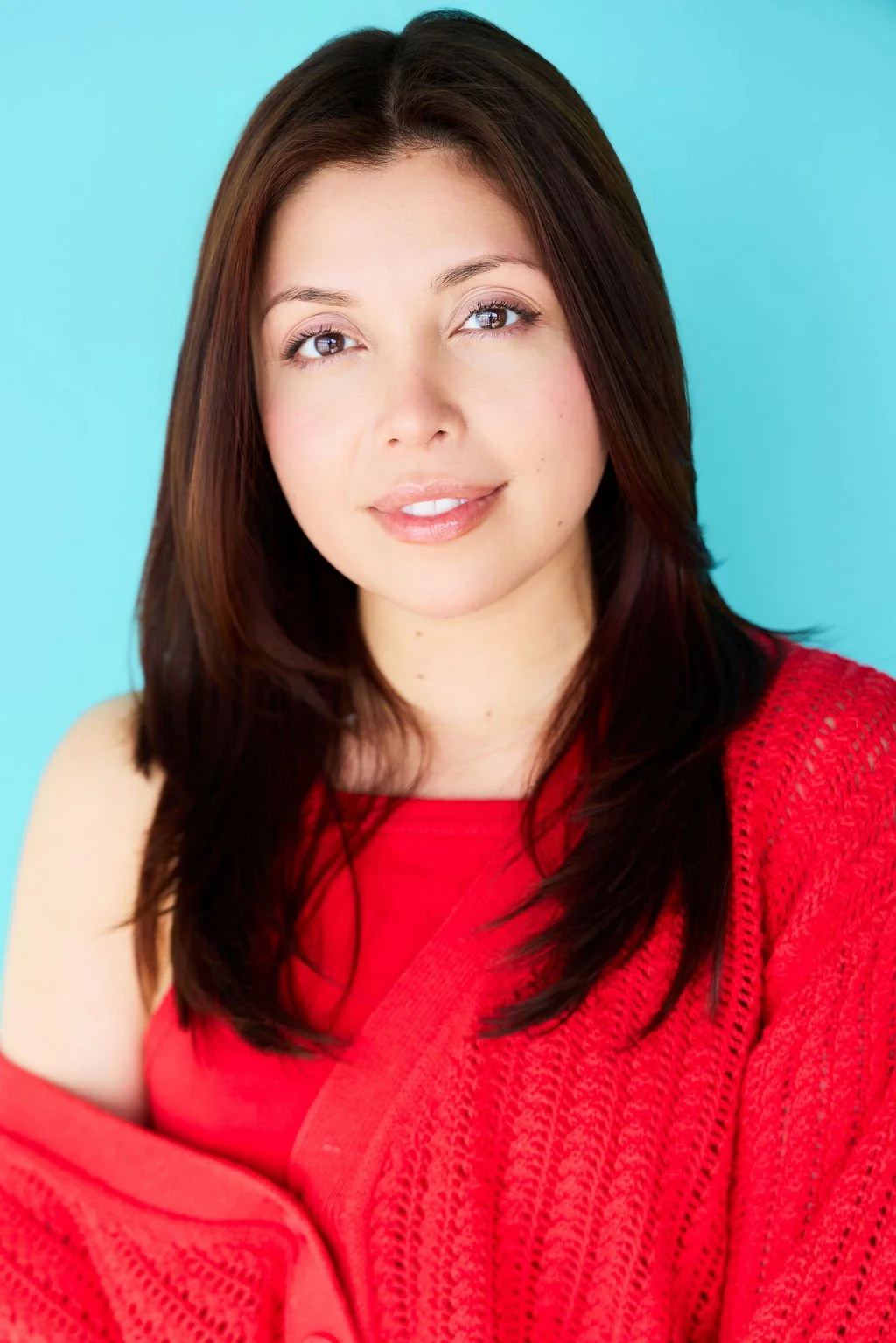 Close-up portrait of a woman with shoulder-length brown hair, light skin, wearing a red knit sweater and a red top, against a light blue background.