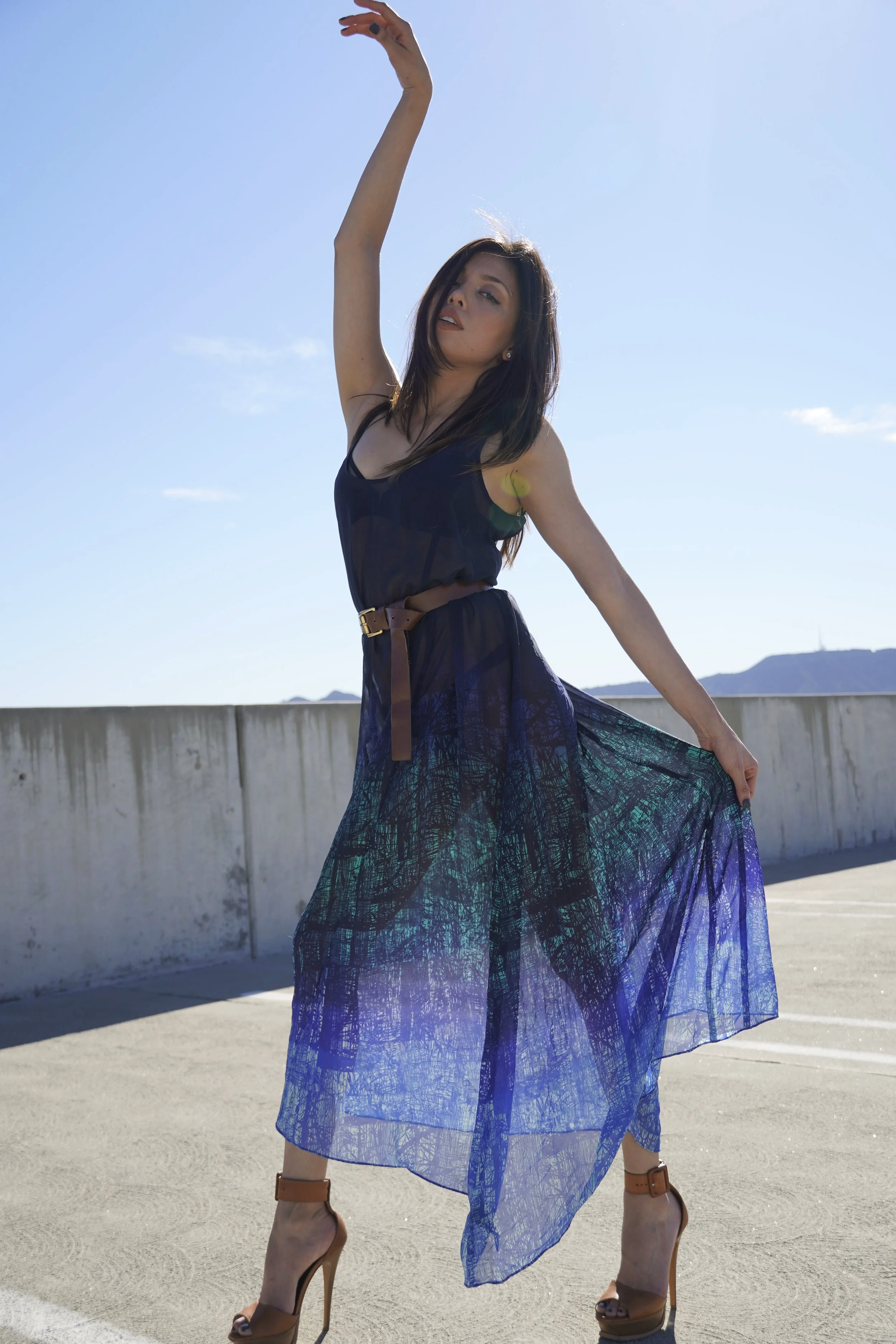 A woman outdoors on a sunny day, wearing a black top and a flowing blue patterned skirt, with her arms raised and head tilted to the side, standing on a concrete surface with mountains in the background.