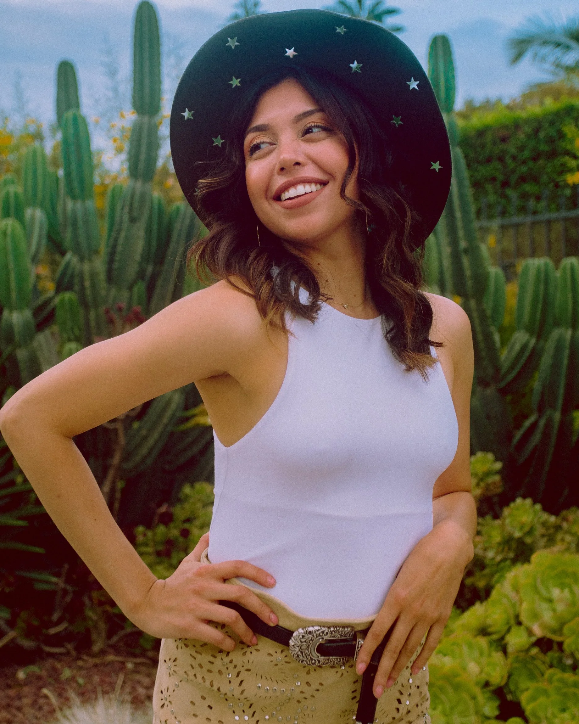 A woman in a cowboy hat decorated with stars, wearing a white sleeveless top, standing outdoors with tall cacti and desert plants in the background.