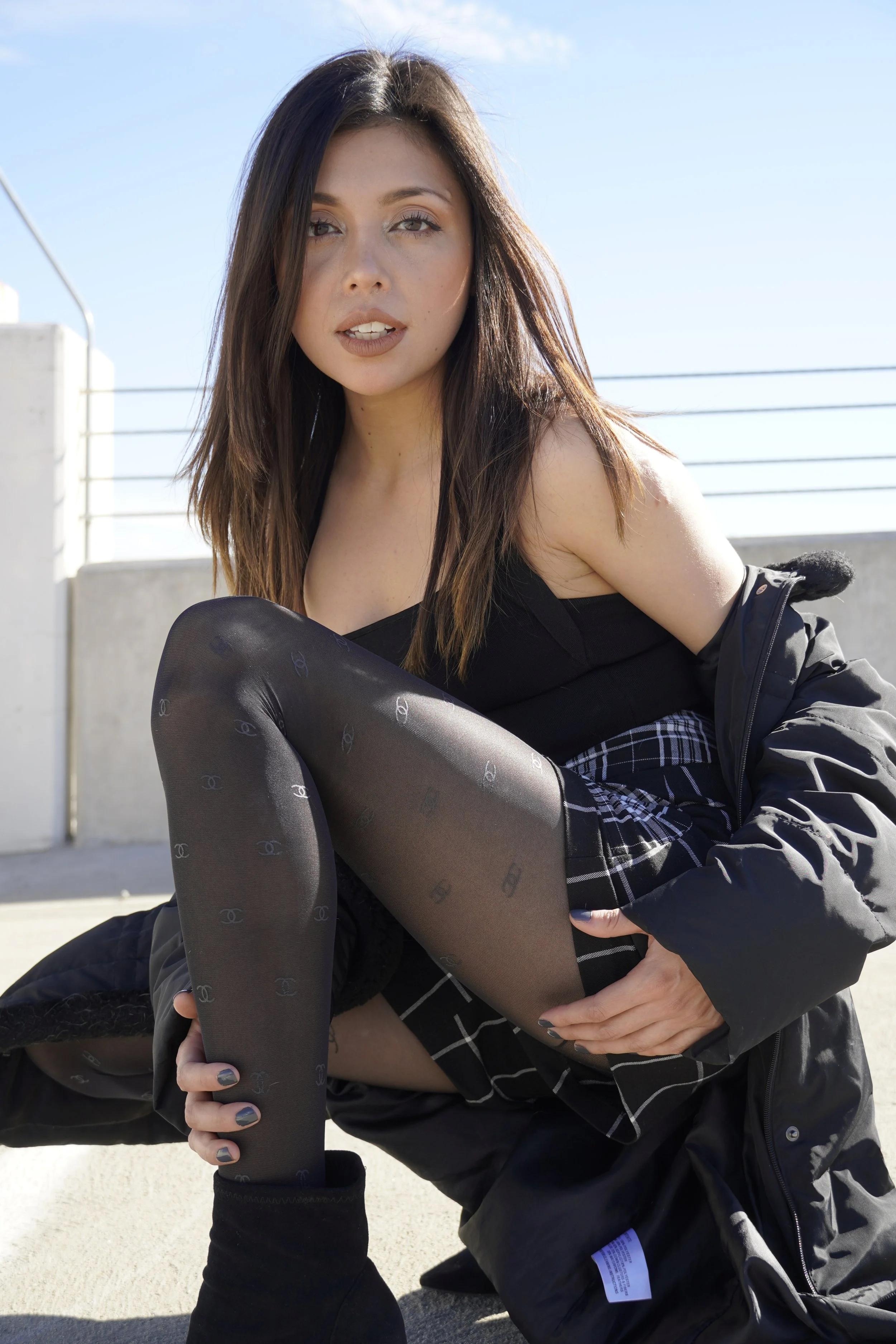 Young woman with shoulder-length brown hair, sitting outdoors on a concrete surface, wearing black tights with a pattern, a black top, and a black jacket, with a blue sky and a railing in the background.