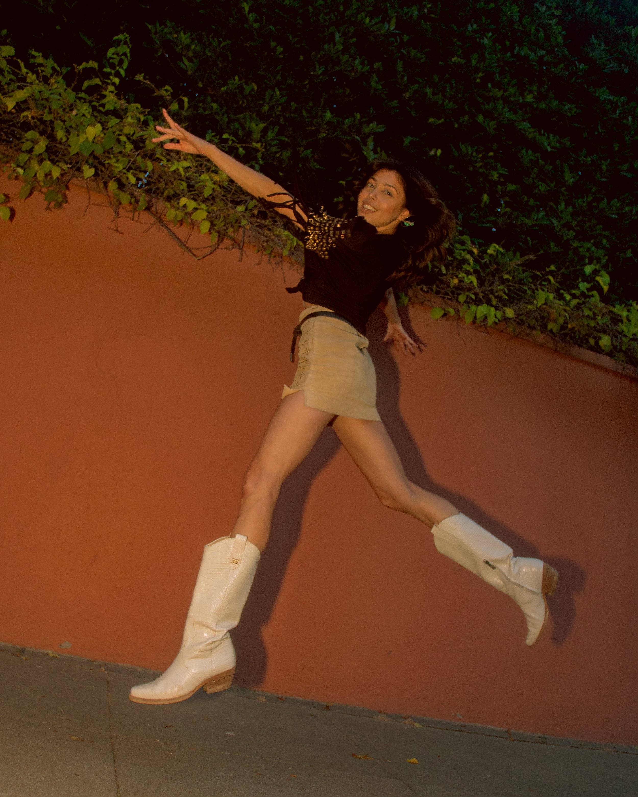 A woman with dark hair, smiling, wearing a black top with gold embellishments on the sleeves, beige shorts, and white boots, happily jumps across the screen in front of an orange wall and dark foliage.