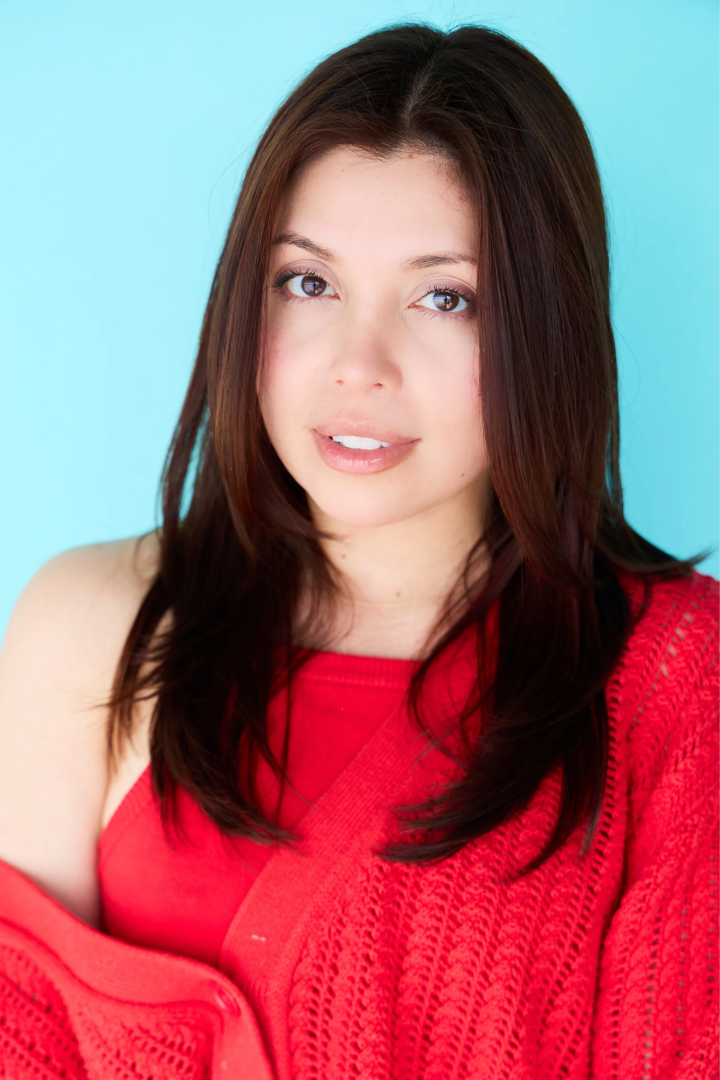 A young woman with long, dark brown hair, light skin, and brown eyes, wearing a red textured sweater, posing against a light blue background and smiling softly.