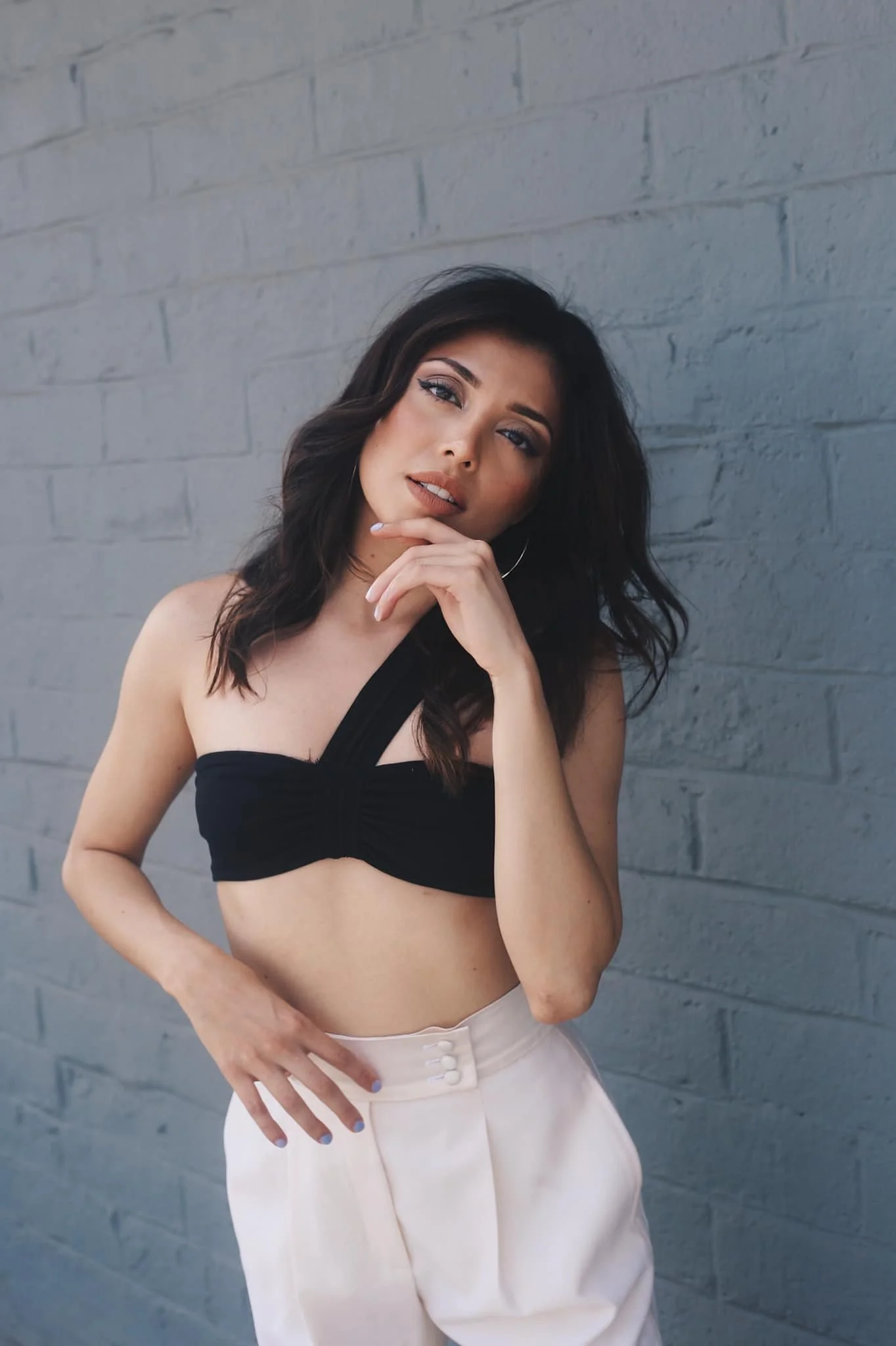 A young woman with dark wavy hair, wearing a black strapless top and light beige high-waisted pants, stands against a gray brick wall, with one hand near her chin and the other near her waist, looking at the camera with a confident expression.