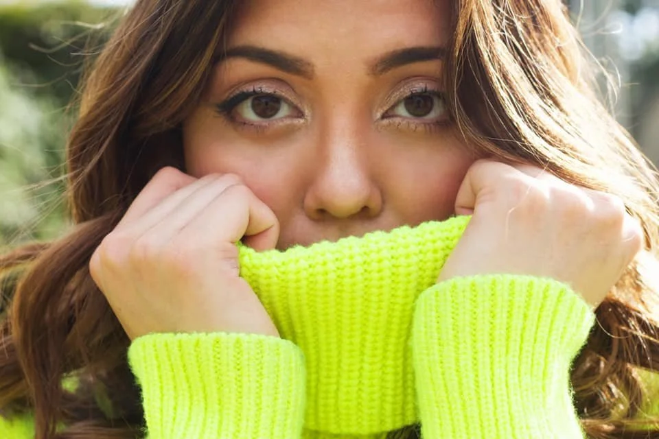 A woman with brown hair and light makeup holding the collar of her neon yellow sweater up to her face.