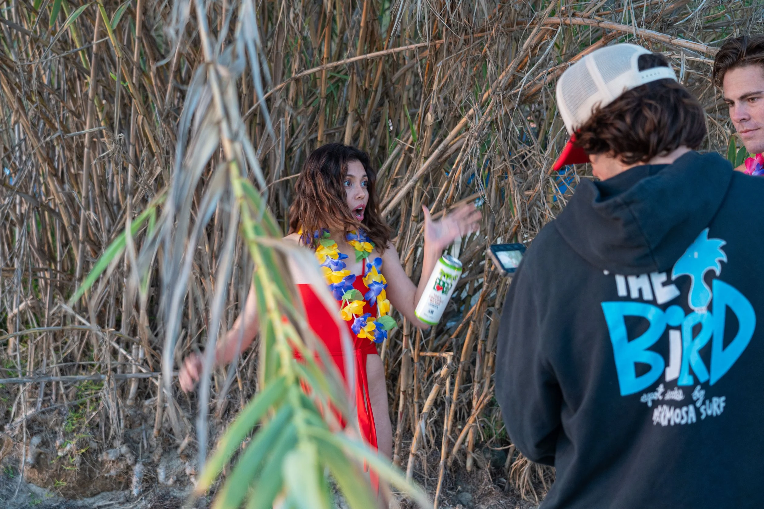 A young woman with long, curly hair wearing a red dress and a yellow flower lei, speaking and gesturing with her hand while speaking to two young men in a dense, dry-branched outdoor setting. One of the men is seen from the back with short curly hair, wearing a black hoodie with blue and white graphics, and a white and red trucker hat. The other man is facing the woman, with short dark hair and a serious expression.