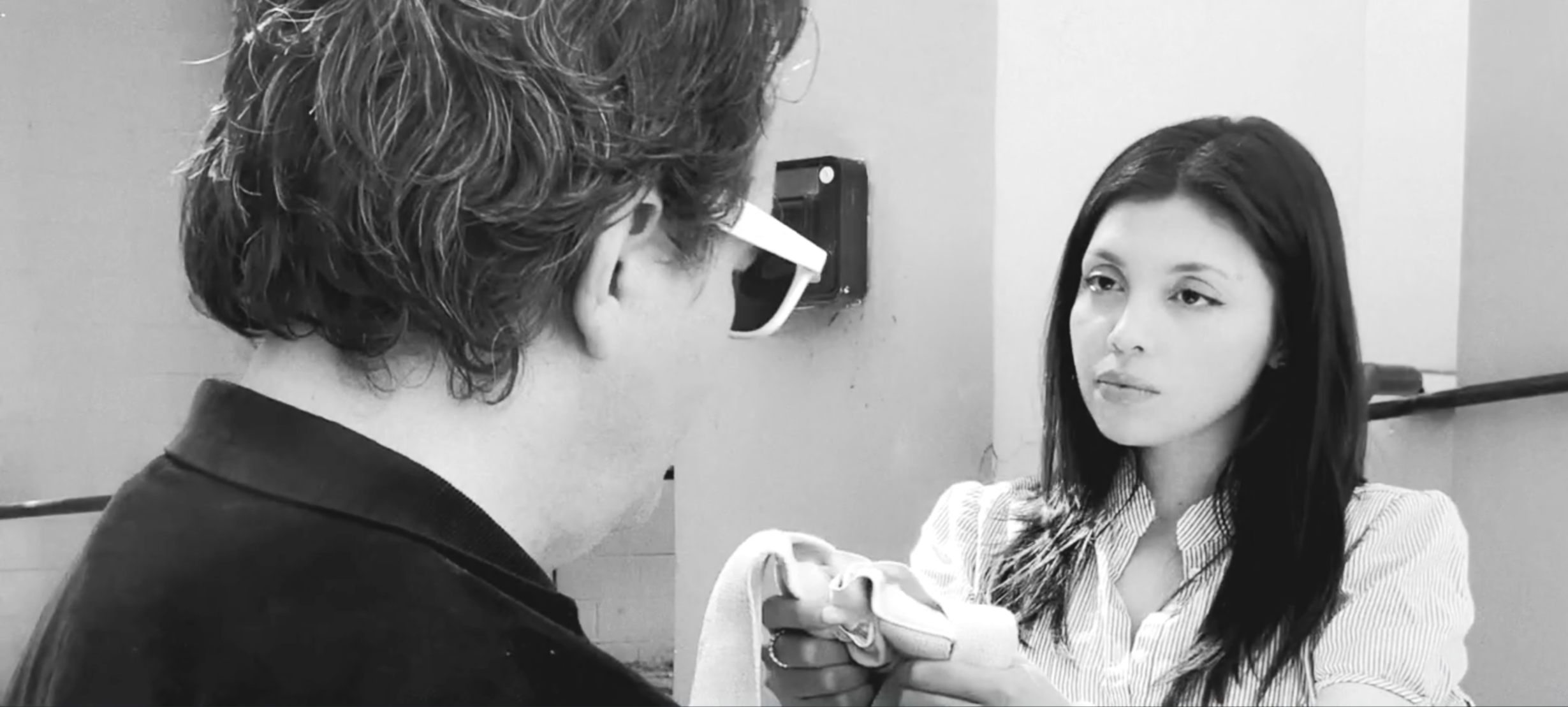A young woman with long dark hair and striped shirt holds a small kitten, engaging with a man with glasses and curly hair, in a room with neutral walls.