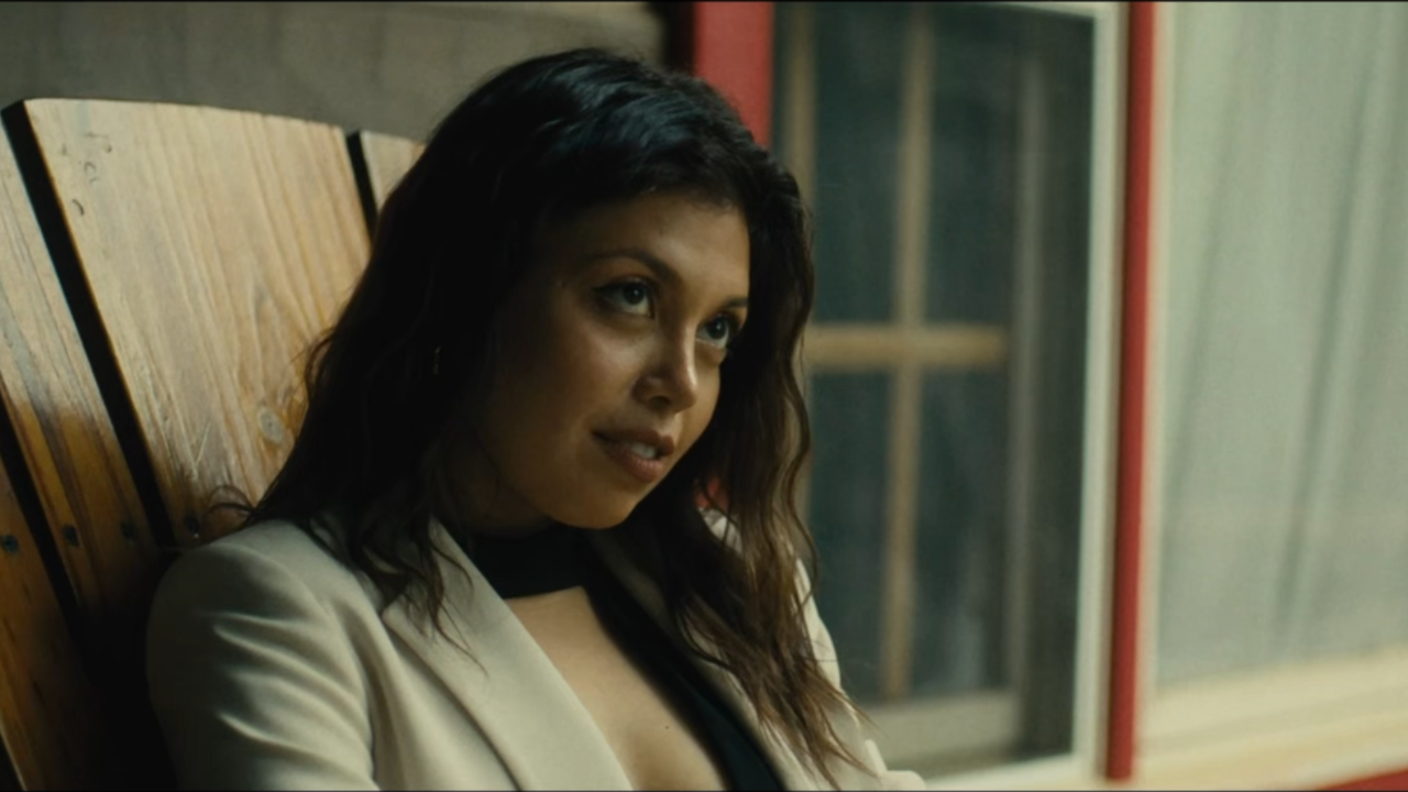 A woman with dark wavy hair sitting on a wooden bench, looking to her left, in an indoor setting with a window and red framing in the background.