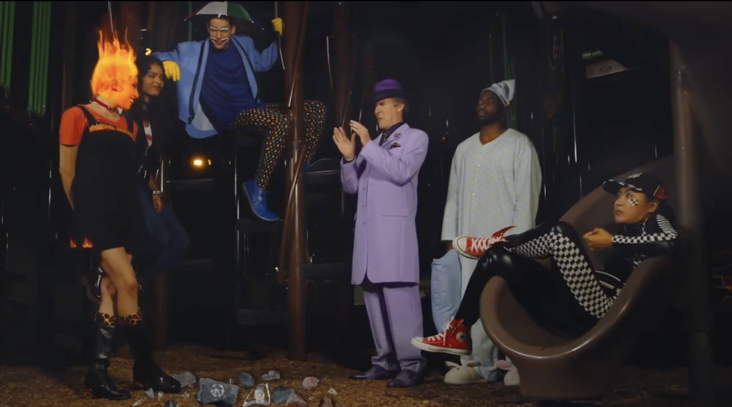 A group of friends at a playground at night. One person has hair on fire, others are standing or sitting nearby, wearing colorful and quirky outfits. The scene has a playful, eclectic vibe.