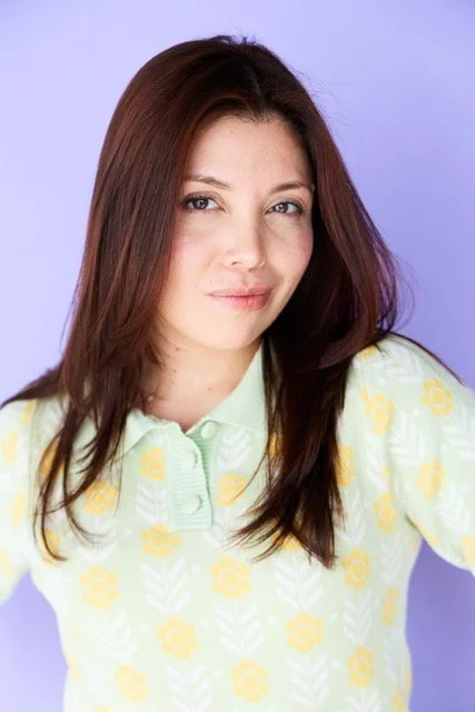 Young woman with shoulder-length dark brown hair, wearing a light yellow shirt with a floral pattern, posing against a light purple background.