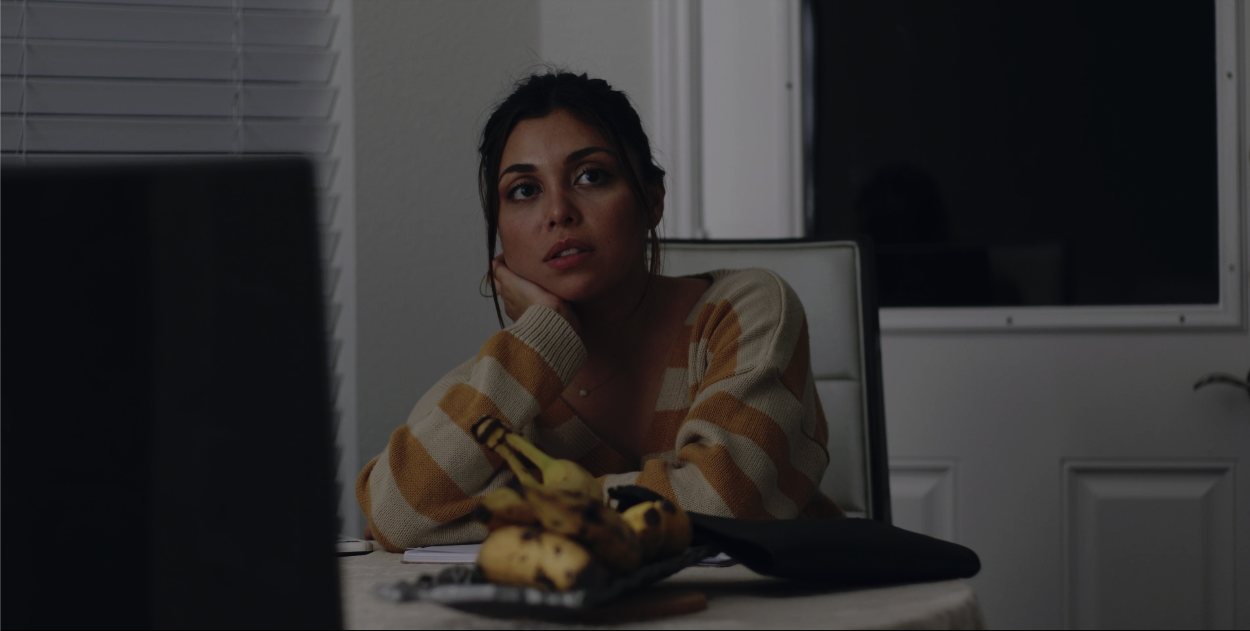 A woman with dark hair and wearing a yellow and beige striped sweater, sitting at a table with her head resting on her hand, appears to be looking at a computer screen in a dimly lit room. There are bananas and papers on the table.