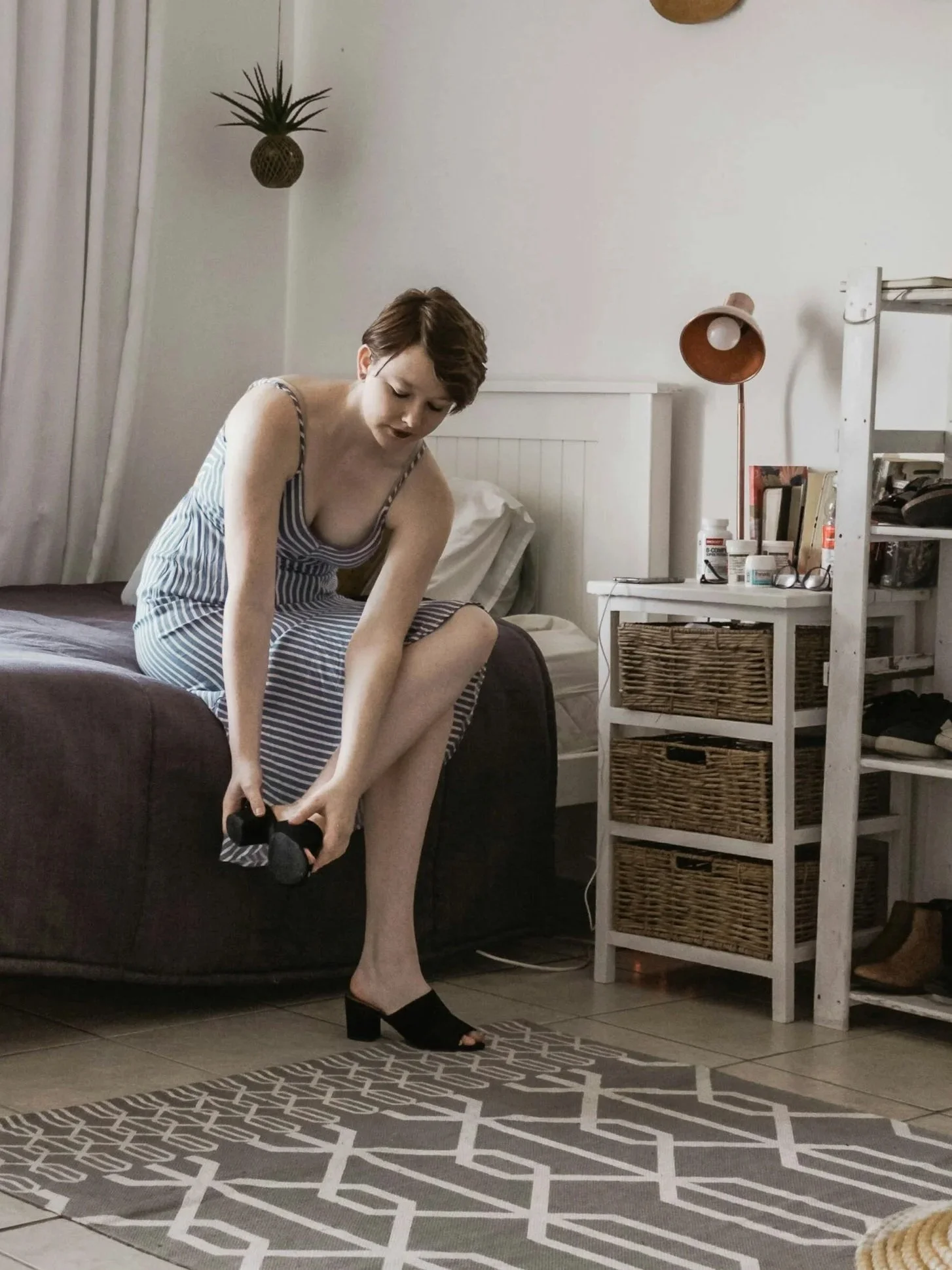 A woman in striped dress and black heeled shoes sitting on a bed, putting on shoes in a modern bedroom with a patterned rug, white furniture, and decorative plants.