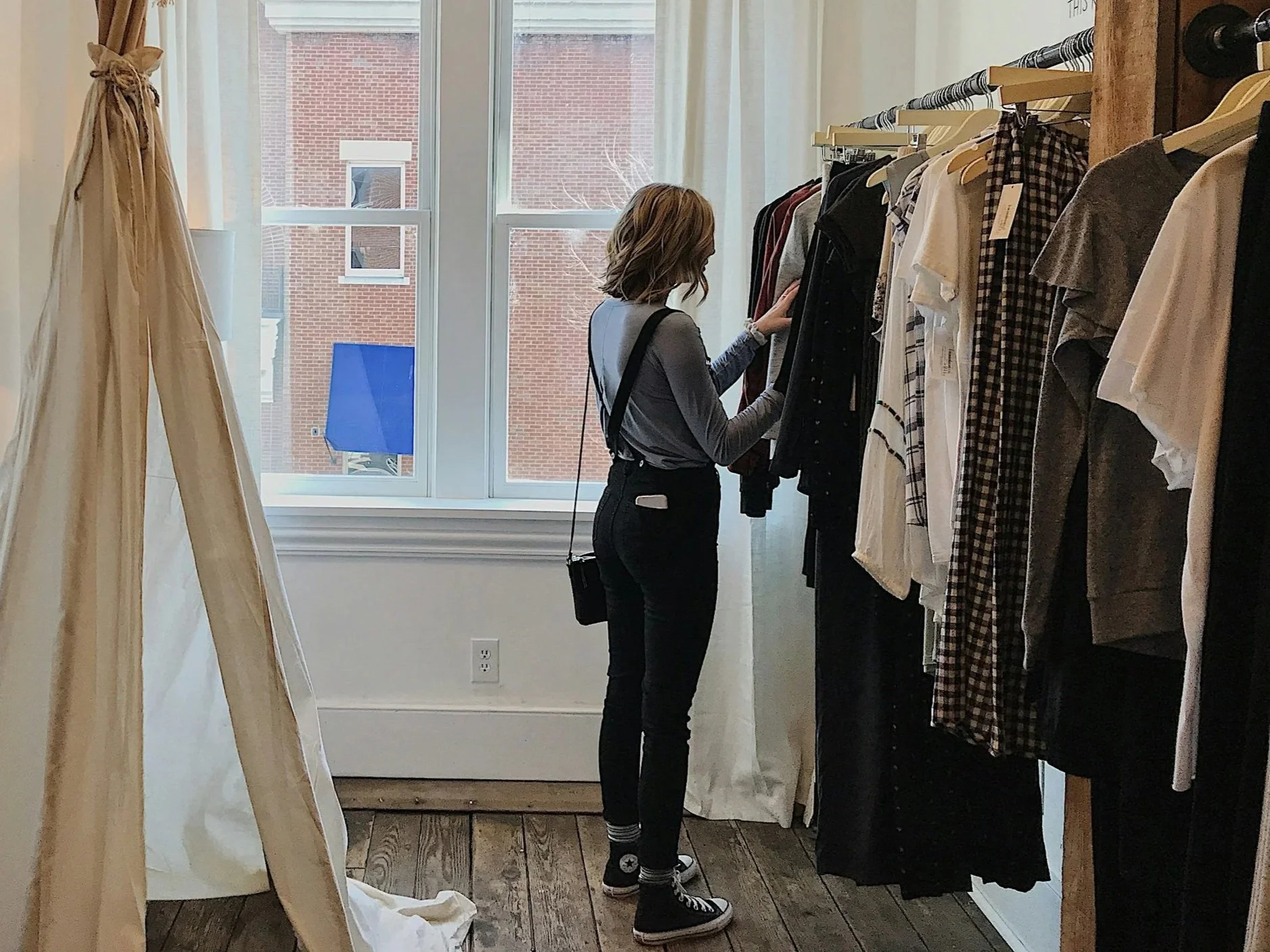 A woman shopping for clothes inside a boutique store, browsing through a rack of casual and checkered shirts, with a large window behind her showing a brick building outside.
