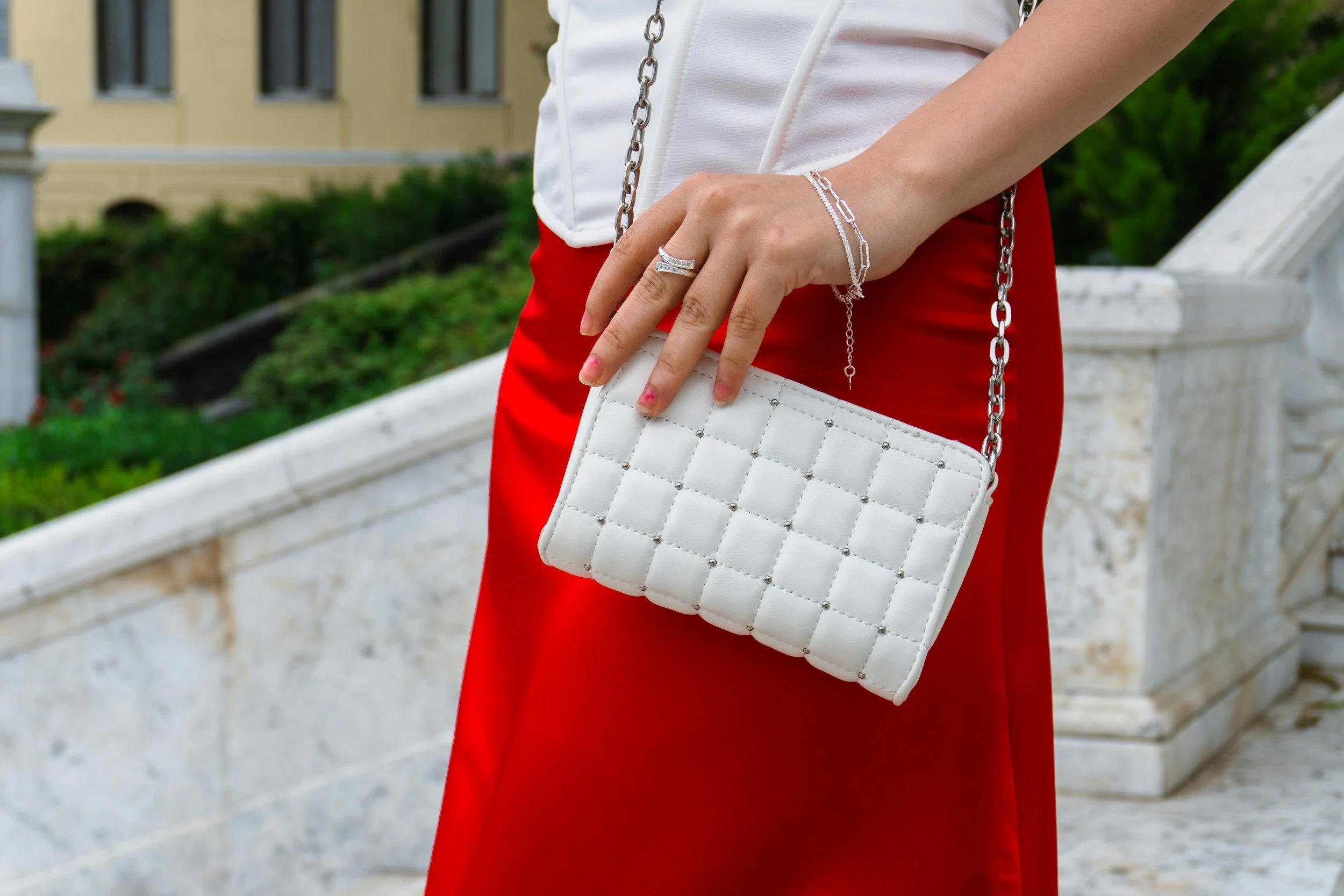 Close-up of a person holding a white quilted handbag with a chain strap, wearing a silver ring and matching silver jewelry on their wrist, next to a white staircase and greenery in the background.