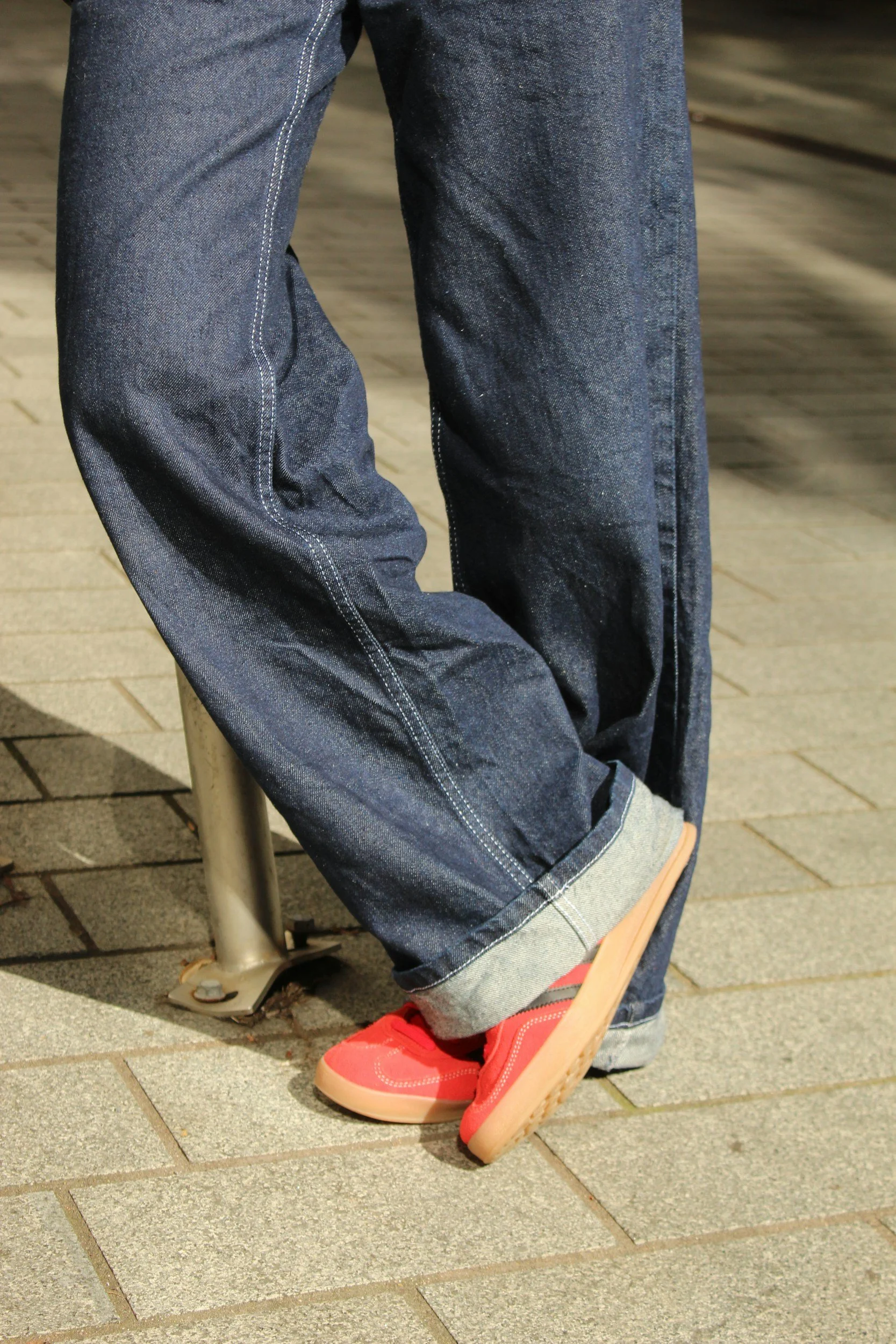 Close-up of a person sitting on a bench, wearing blue jeans and red sneakers, with part of a wooden object visible on the ground.