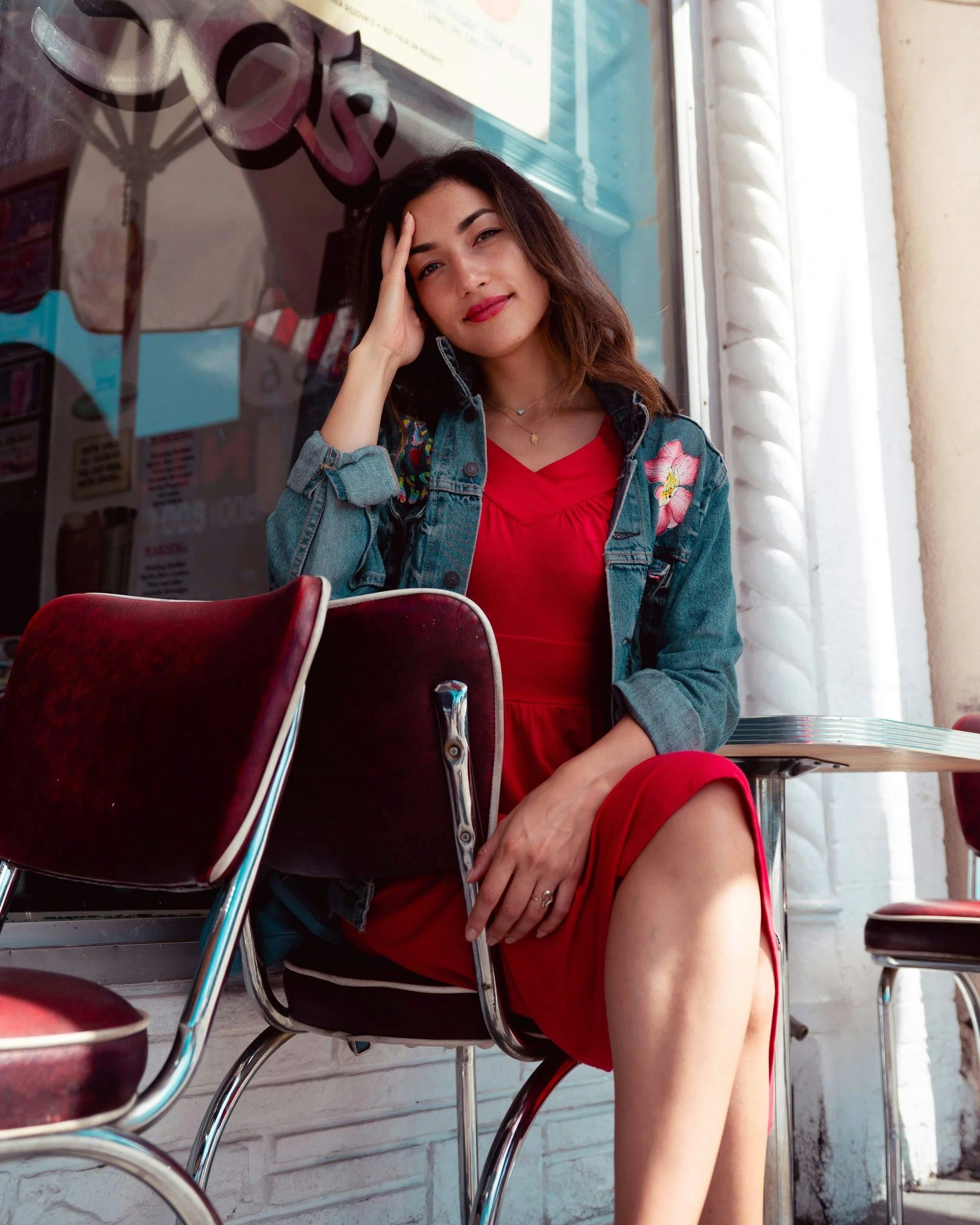 Young woman with brown hair and red lipstick sitting in a diner or cafe. She is wearing a red dress and a denim jacket with floral embroidery. She is resting her head on her hand and looks at the camera with a slight smile. The background includes a white brick wall and some restaurant chairs.