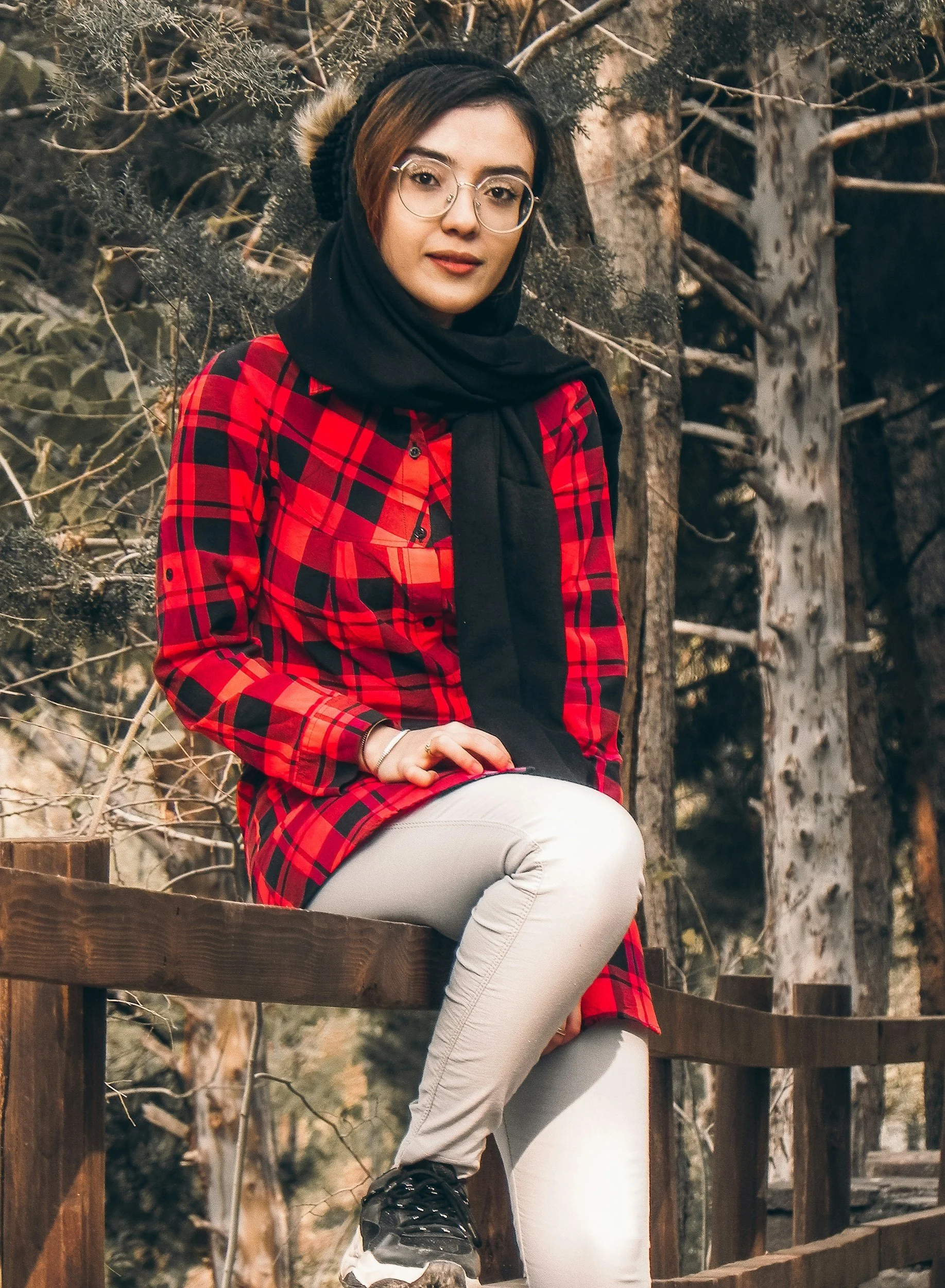 A young woman with glasses and short brown hair, wearing a black beanie and a black scarf, sitting on a wooden railing outdoors. She is dressed in a red plaid shirt, white pants, and black sneakers, with hands resting on her lap. The background features trees and natural scenery.