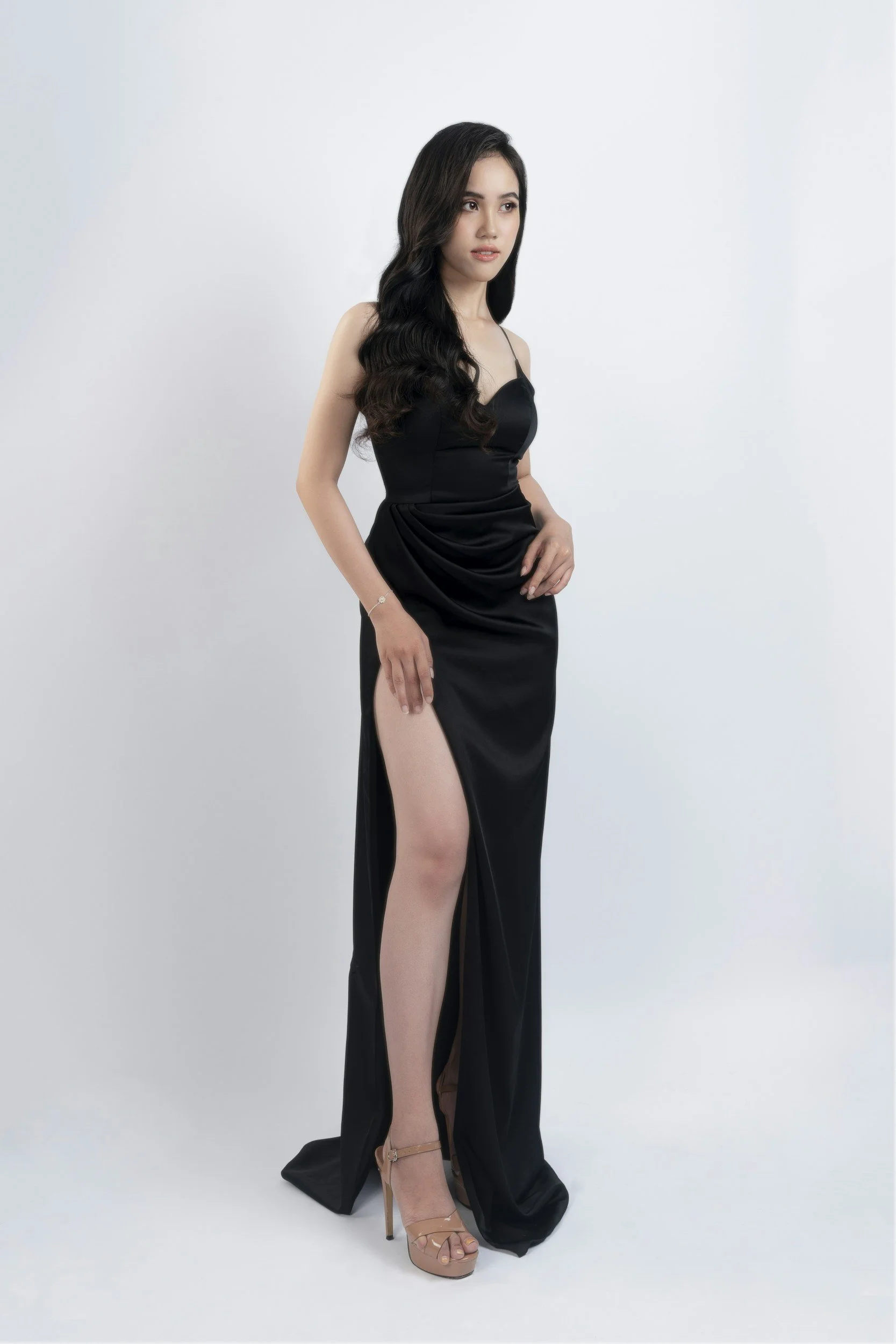 A woman with long dark wavy hair wearing a black dress with a high slit, beige high heels, standing against a plain white background.