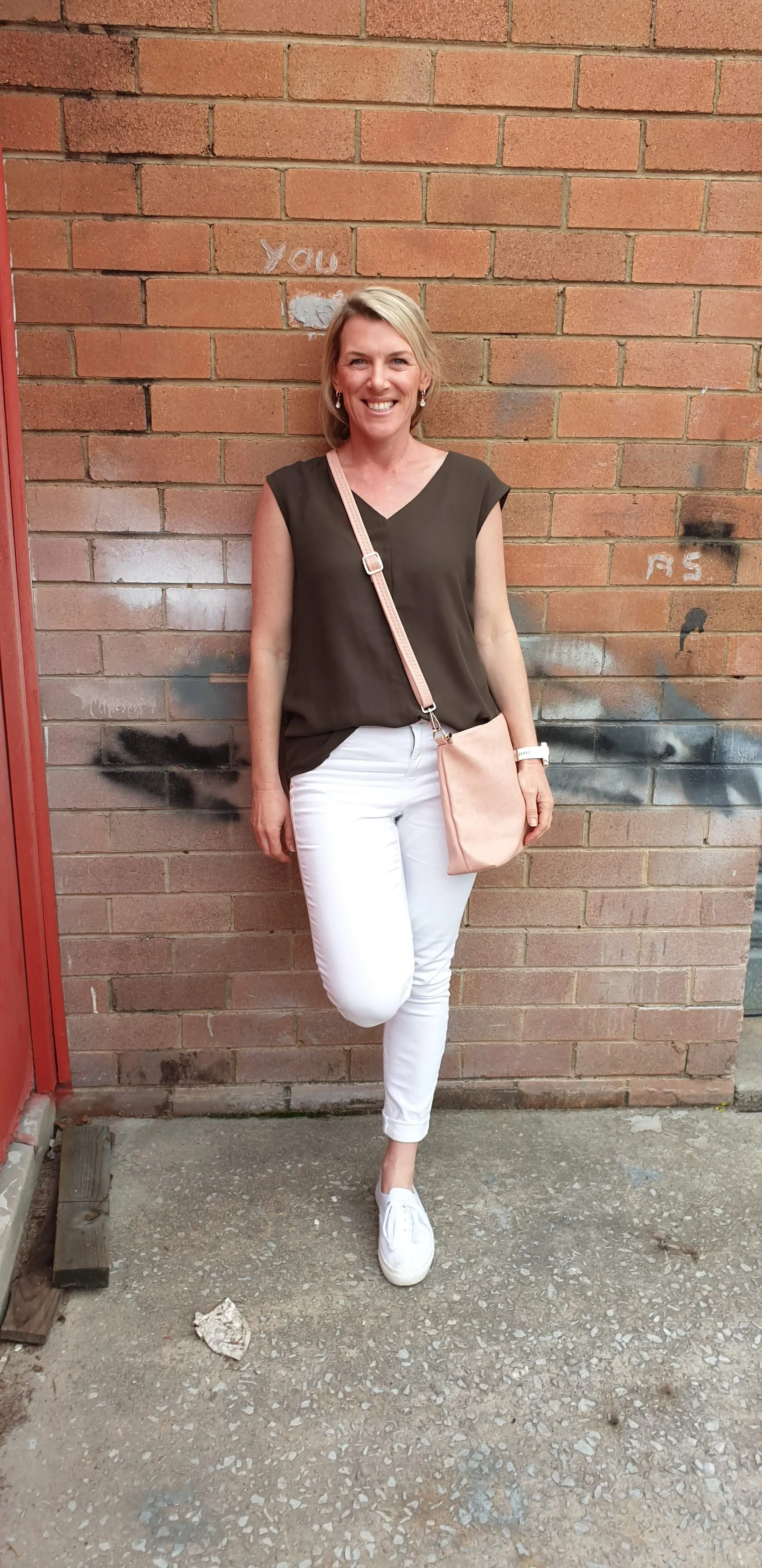 A woman standing against a brick wall, smiling, wearing a sleeveless brown top, white pants, white sneakers, and carrying a pink shoulder bag.