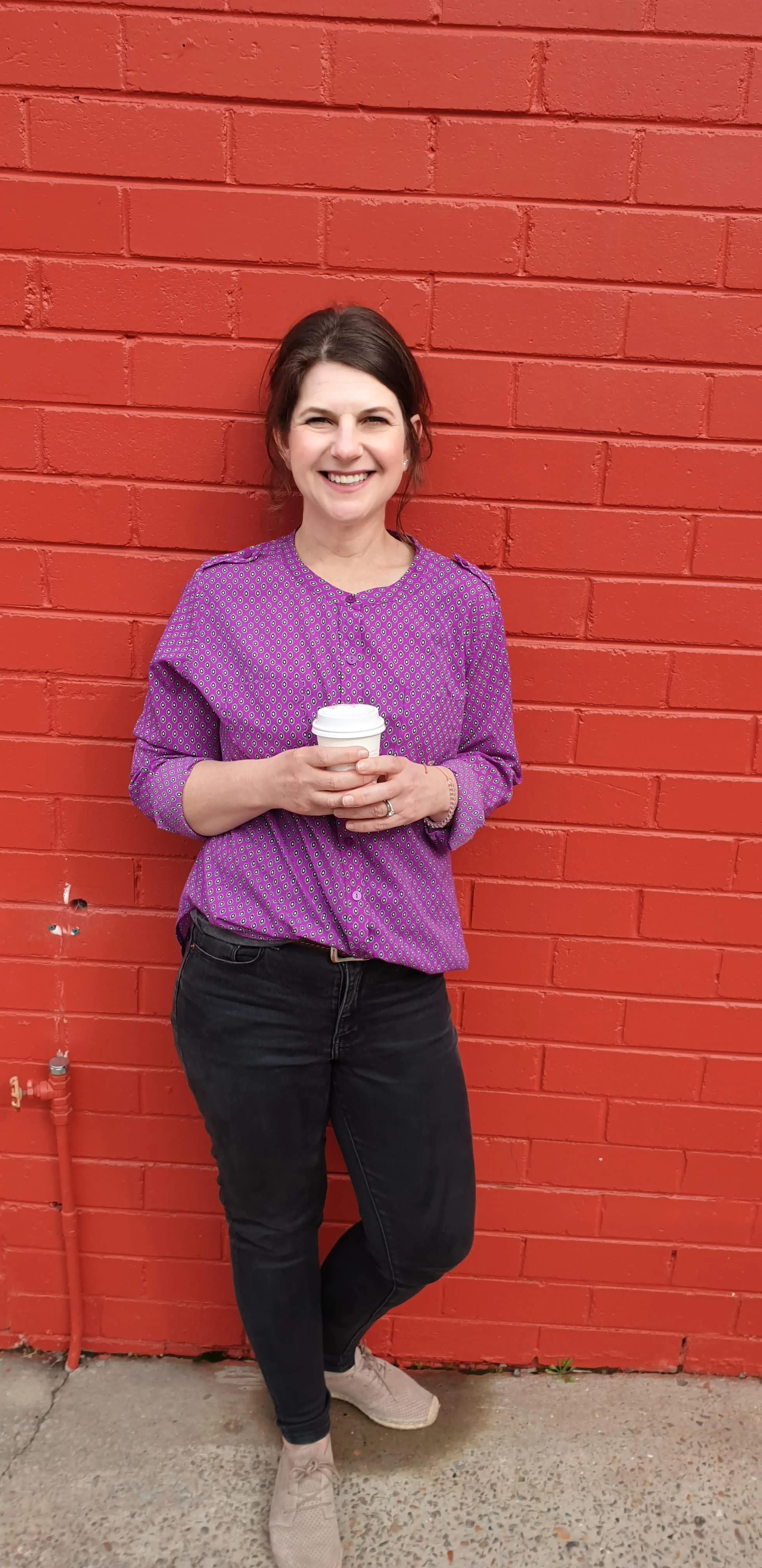 A woman with dark hair in a ponytail, wearing a purple patterned blouse, black jeans, and beige sneakers, standing against a red brick wall, holding a white take-out coffee cup, smiling.