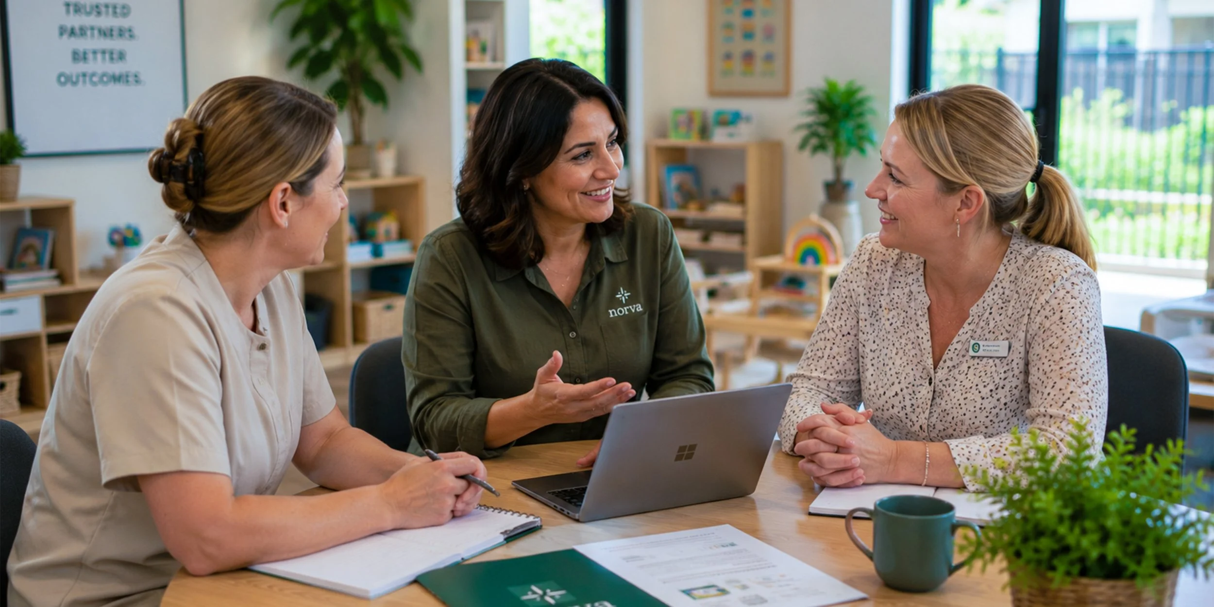 Three women sitting at a table in a bright room, chatting and smiling. One woman is in a beige uniform, another is in an olive green shirt with a 'norva' logo, and the third is in a beige patterned shirt. There's a laptop, notebooks, and a green plant on the table.